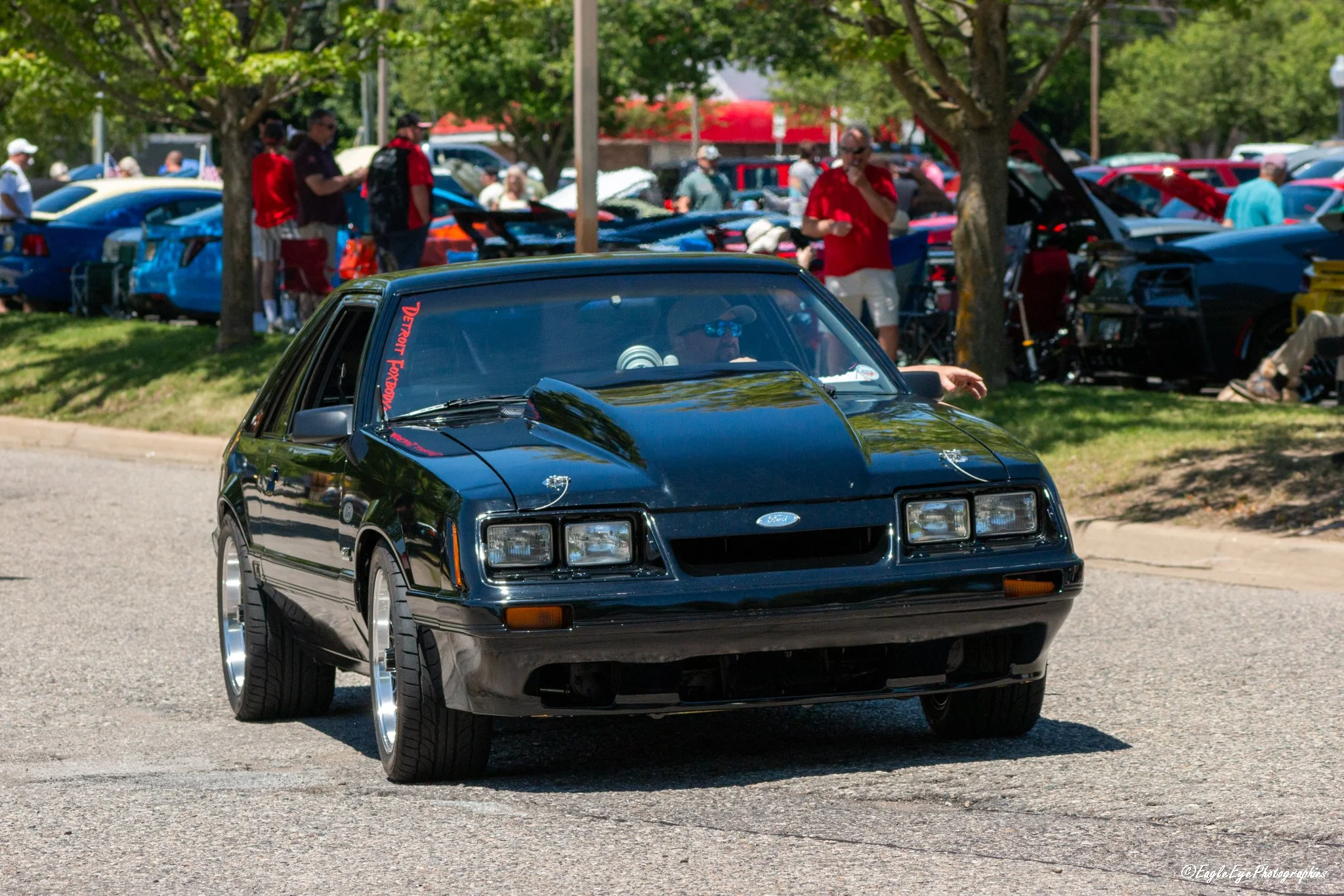 A black sports car in the foreground driving on the street during a car show, with a crowd of people and rows of colorful cars and cars with open hoods in the background.
