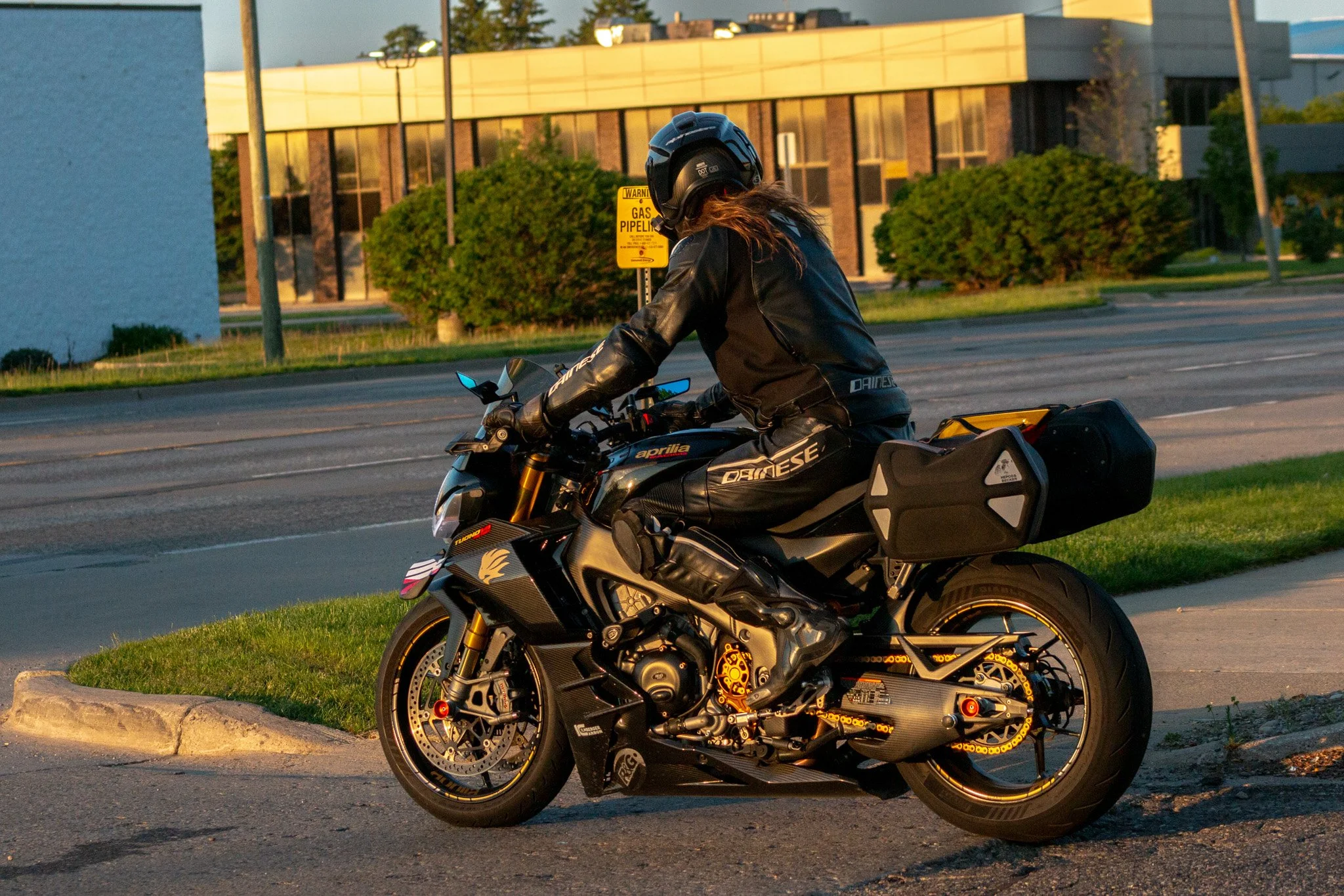 A person wearing a black motorcycle helmet and black leather gear riding a black sport motorcycle on a city street during sunset with a modern office building and green bushes in the background.