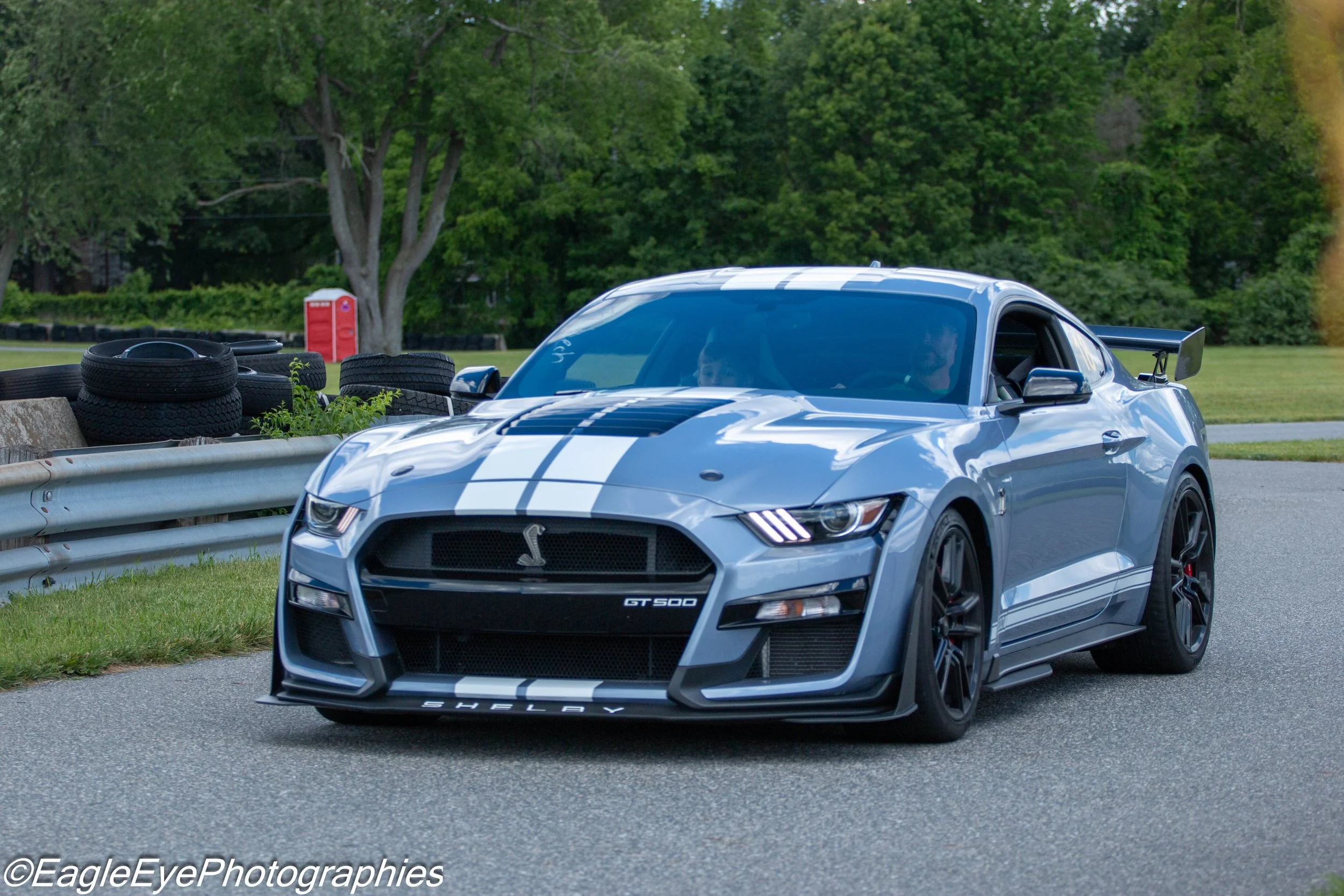 A silver Ford Shelby GT500 sports car with black racing stripes parked on the side of a road with green trees in the background. There are stacked tires behind a guardrail in the scene.