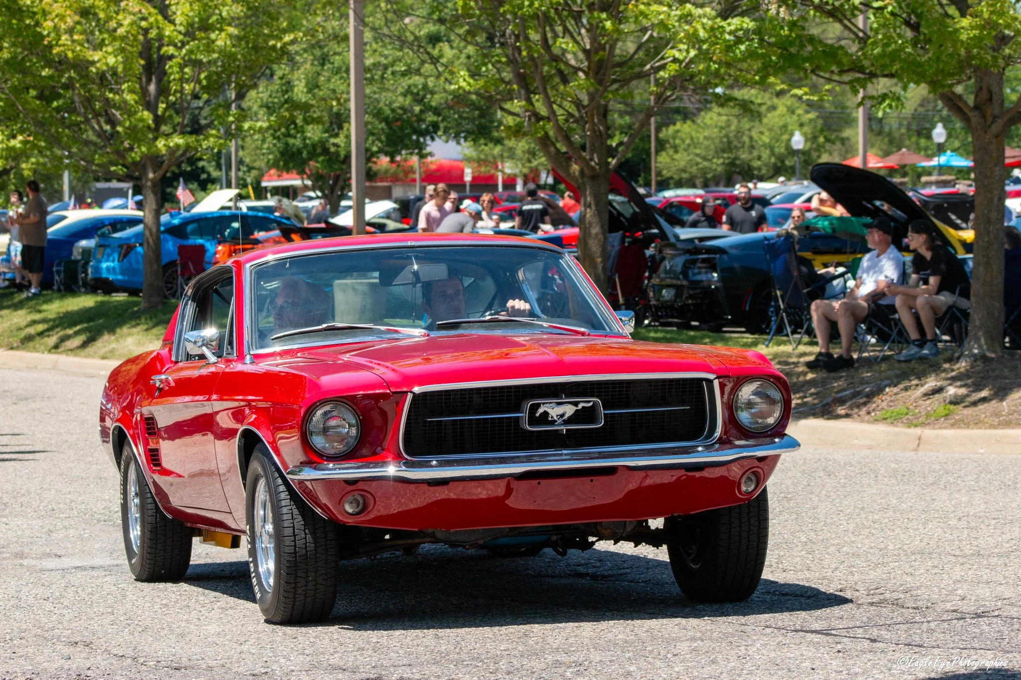 Red classic Ford Mustang driving through a car show with people and other cars in the background, some with open hoods, under green trees on a sunny day.
