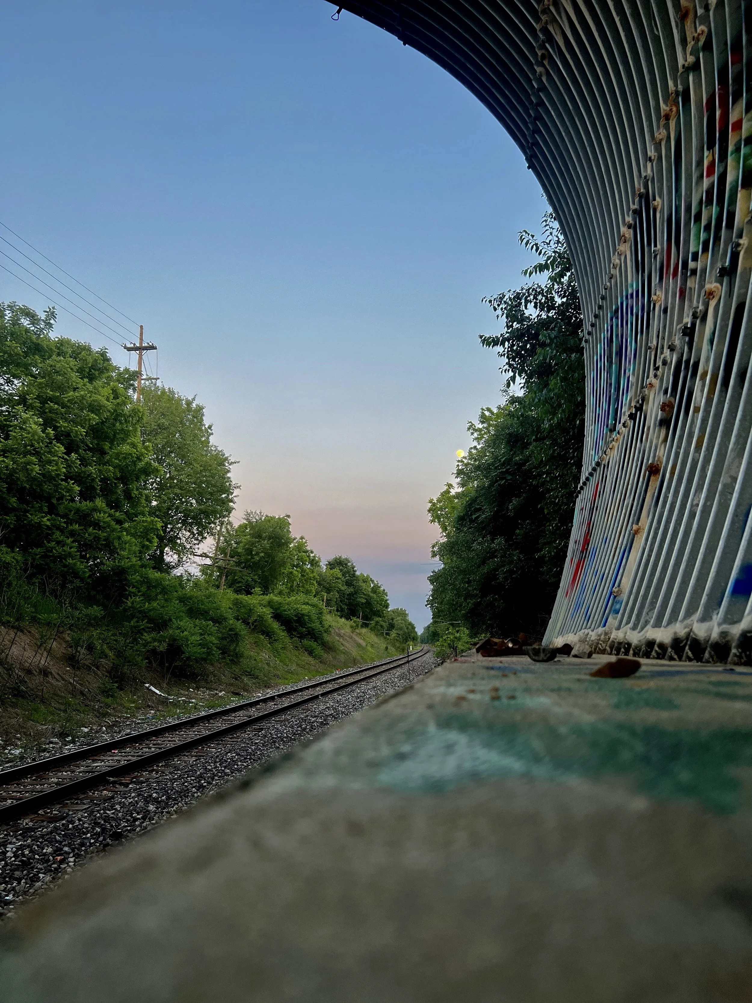 View of railroad tracks running through a green area with trees on both sides, seen from a low angle near a metallic structure on the right, with a clear blue sky overhead and some power lines in the distance.