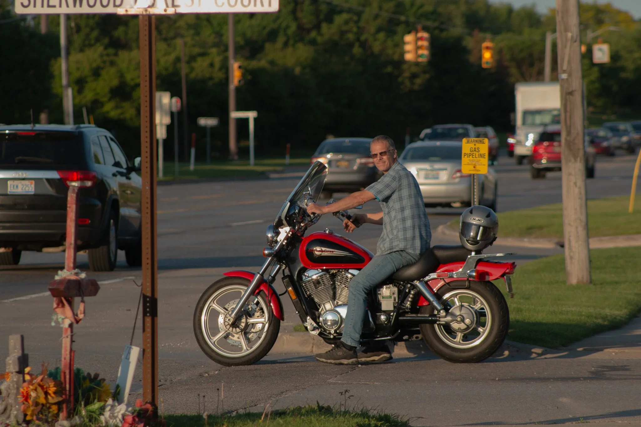 A man riding a red motorcycle on a street with cars in the background, a street sign for Sherwood Drive, and a warning sign for a gas pipeline.