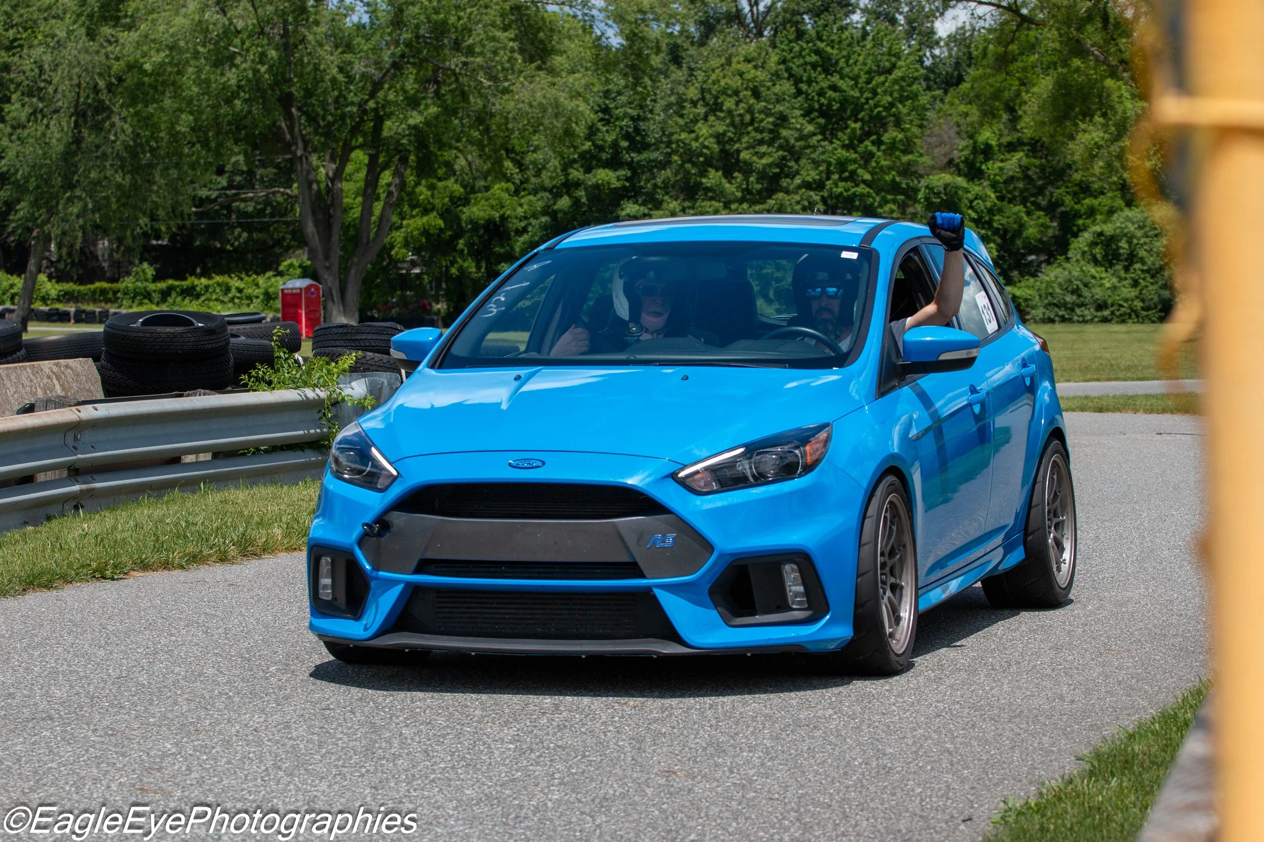 A blue Ford Fiesta RS race car with a driver and a passenger wearing helmets, parked near a guardrail with tires stacked behind it. The background features green trees and a red mailbox.