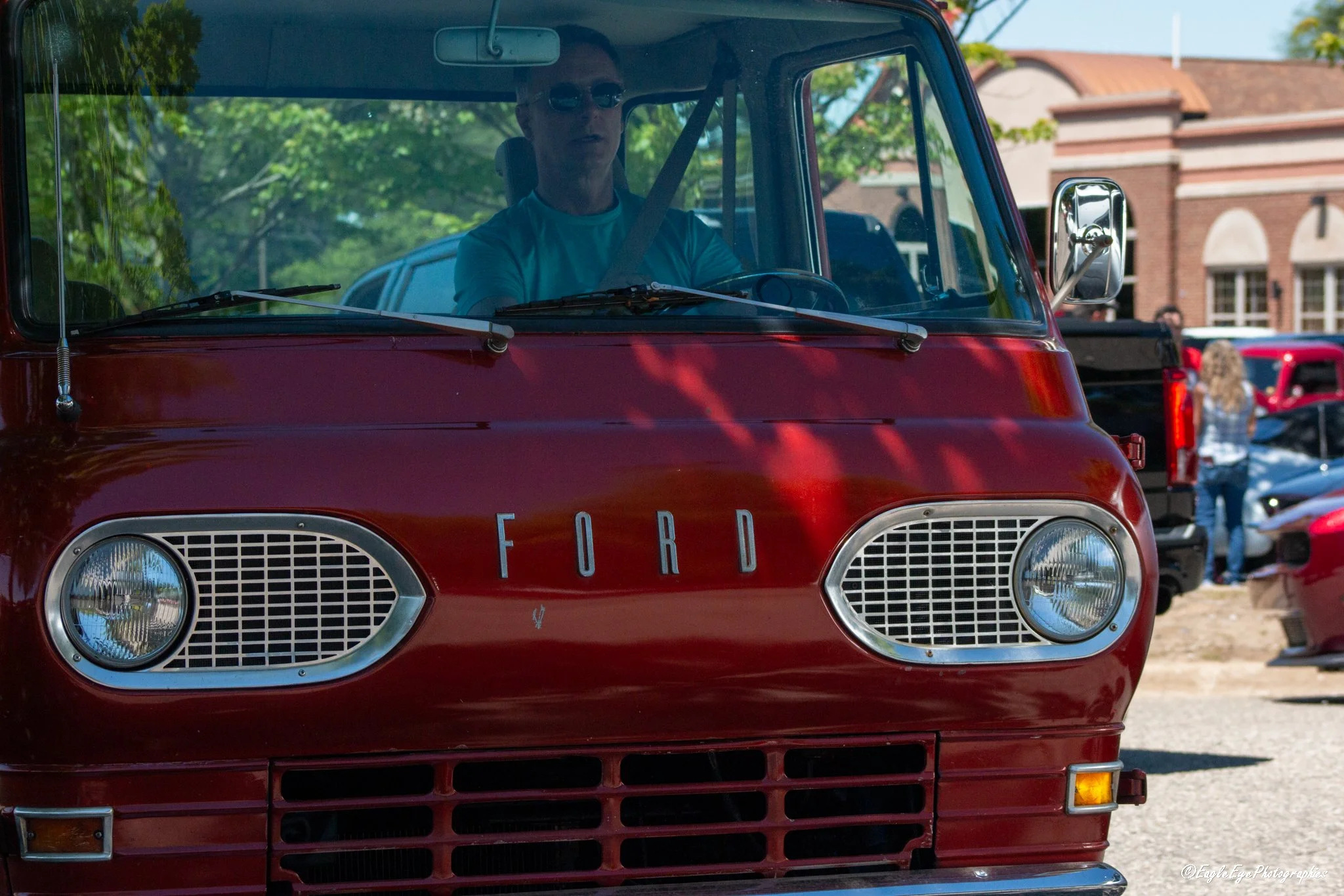 Front view of a vintage red Ford truck with a person inside wearing sunglasses, parked outdoors at an event with other vehicles and people visible in the background.