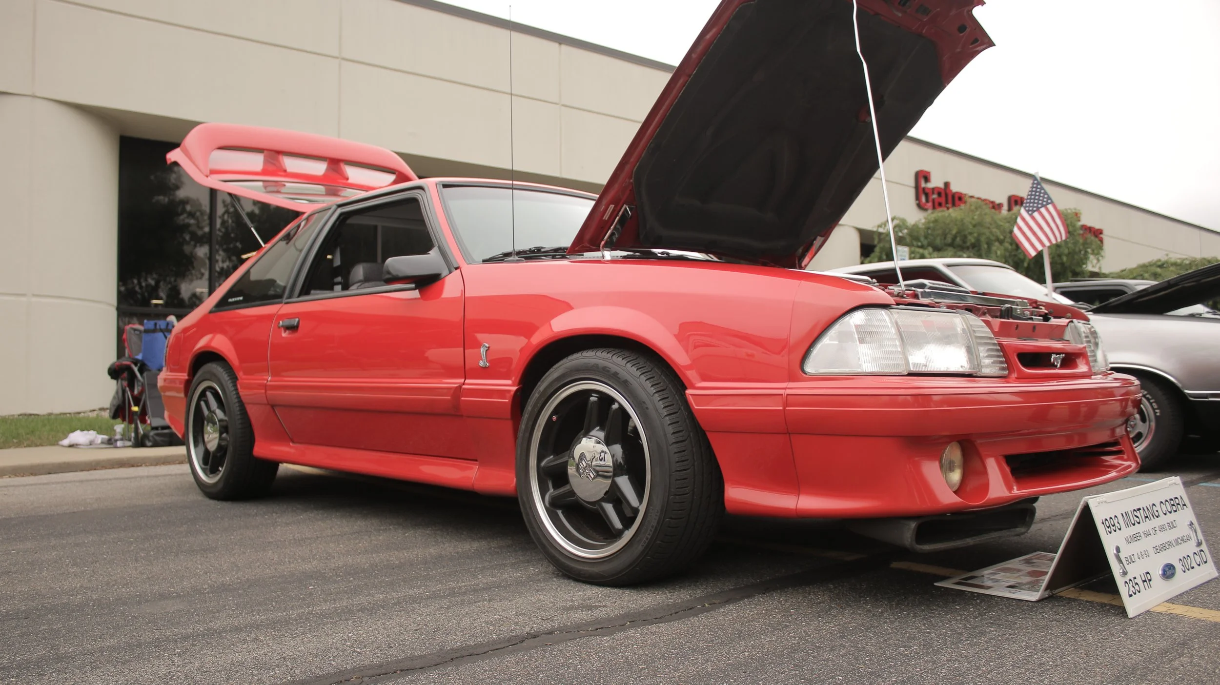 Red 1993 Ford Mustang Cobra on display at a car show with open hood, black wheels, and an American flag on a pole in the background.