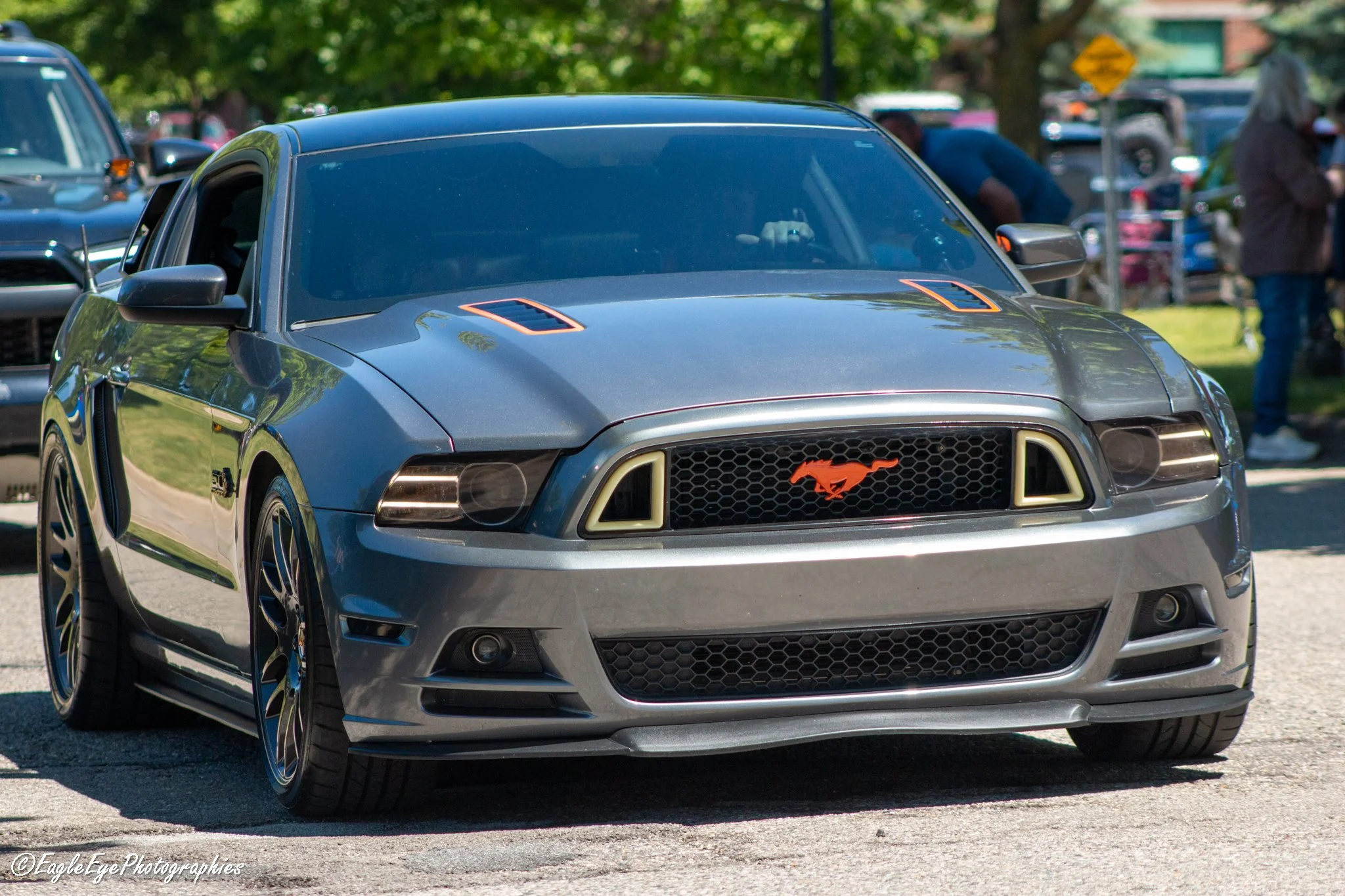 A gray Ford Mustang with a black hood and orange accents, parked outdoors among trees and other cars.