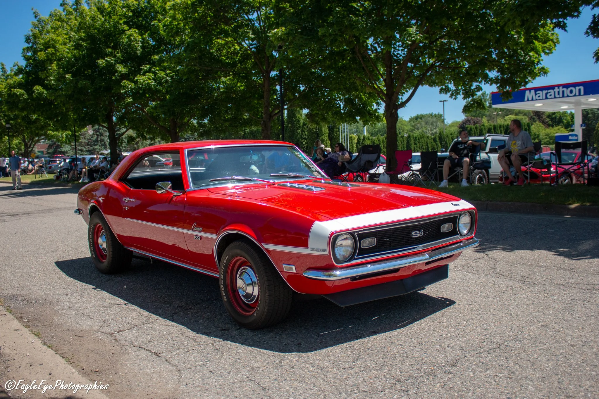 A red classic Chevrolet Camaro SS car at a car show, with people sitting on chairs and a Marathon gas station sign in the background, surrounded by green trees and a sunny sky.