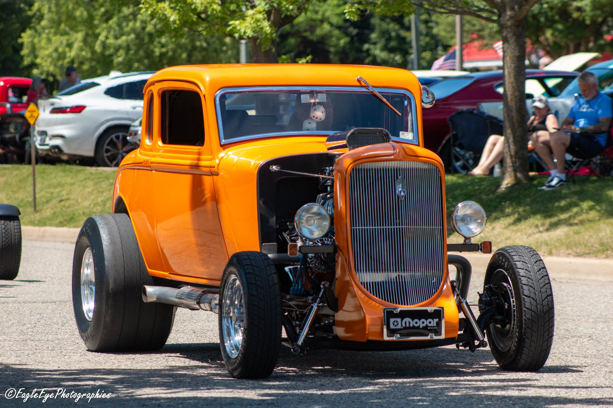 A vintage orange hot rod car with its hood removed on display at an outdoor car show, with other cars and people in the background.