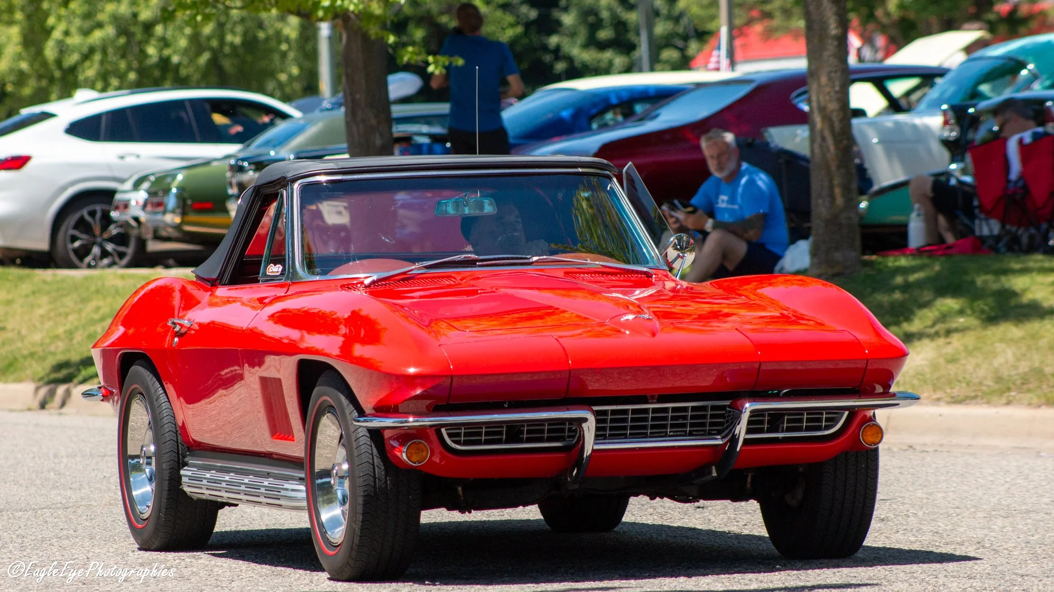 A red vintage Chevrolet Corvette parked at a car show with other cars and people in the background.