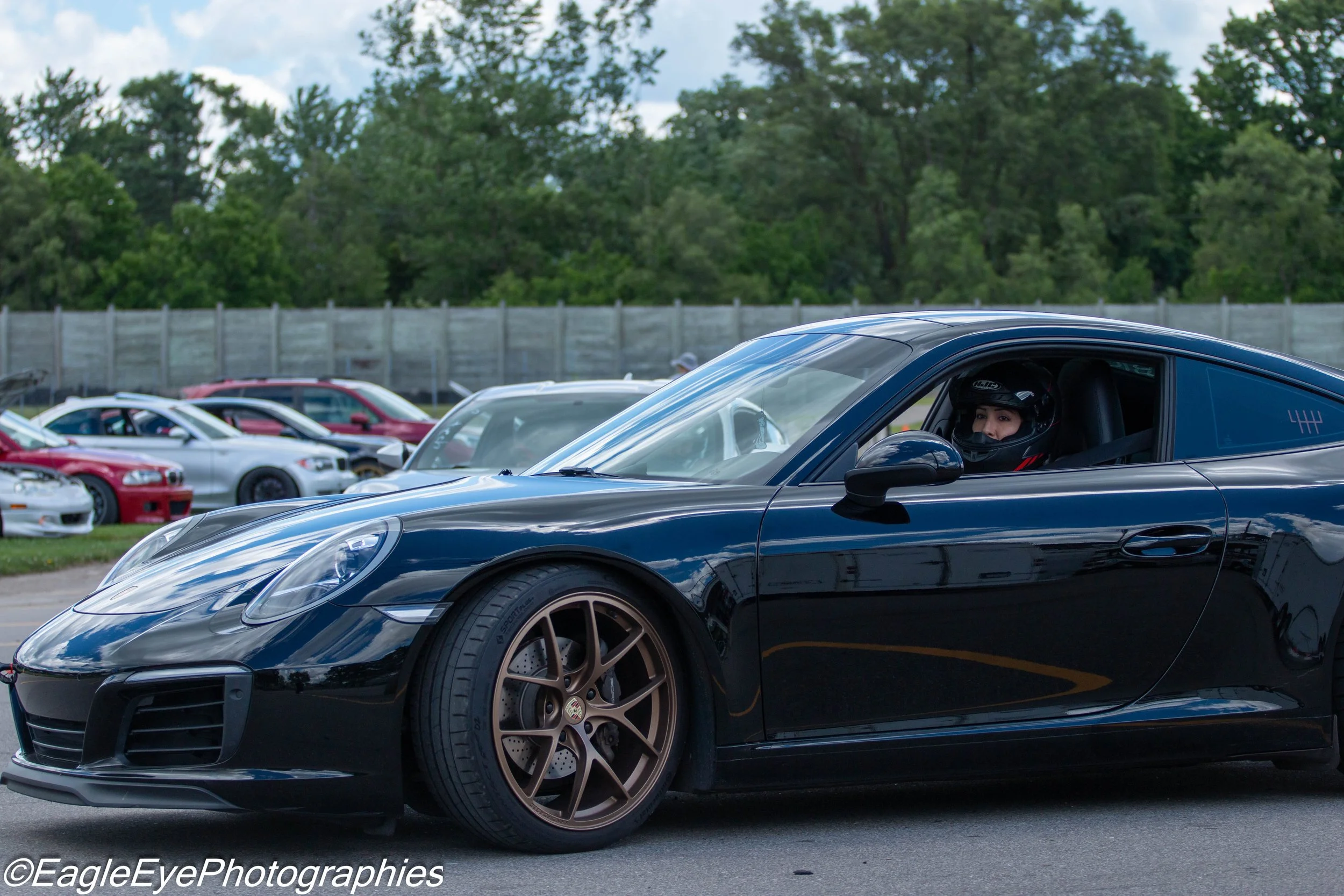 A black sports car with a woman inside wearing a racing helmet at a car meet, with other cars parked behind and a wooden fence with trees in the background.
