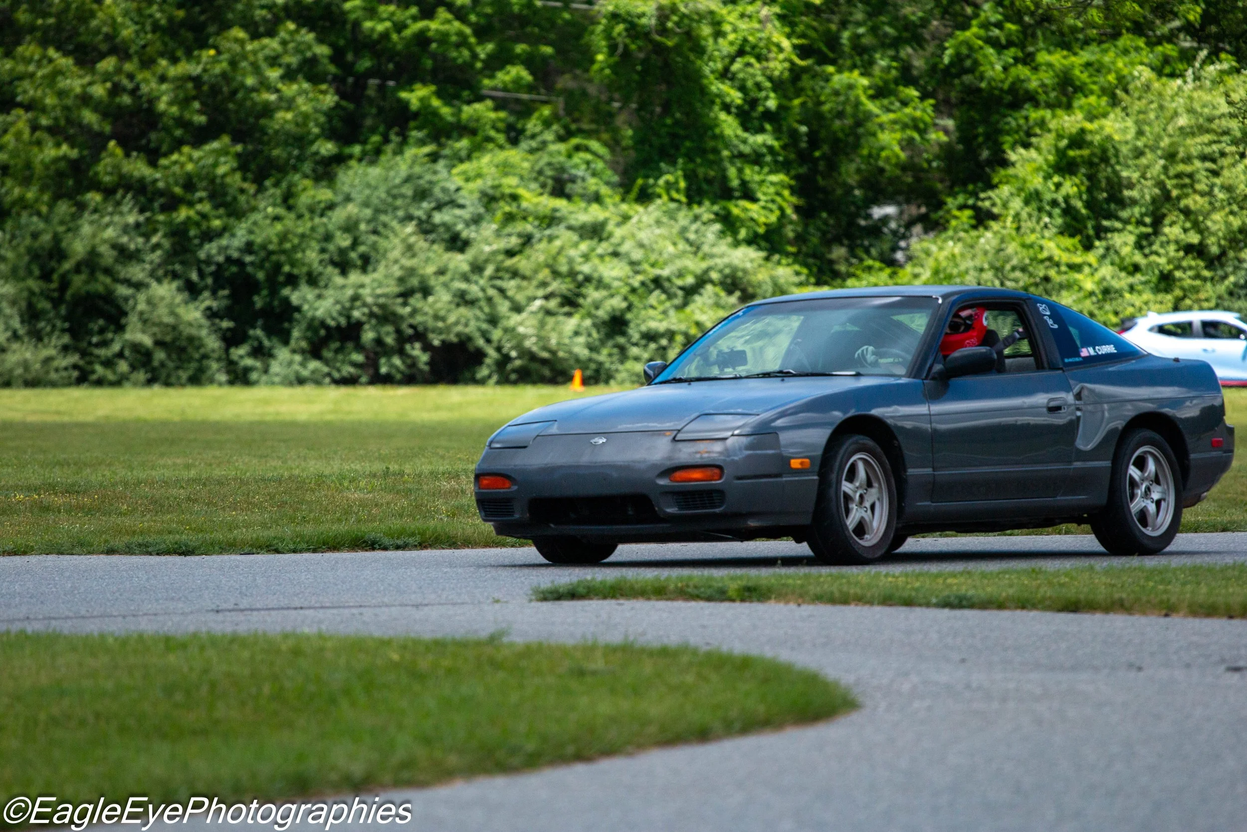 A dark gray sports car parked on a grassy area near a paved street with trees in the background. A person wearing a red helmet is inside the car.