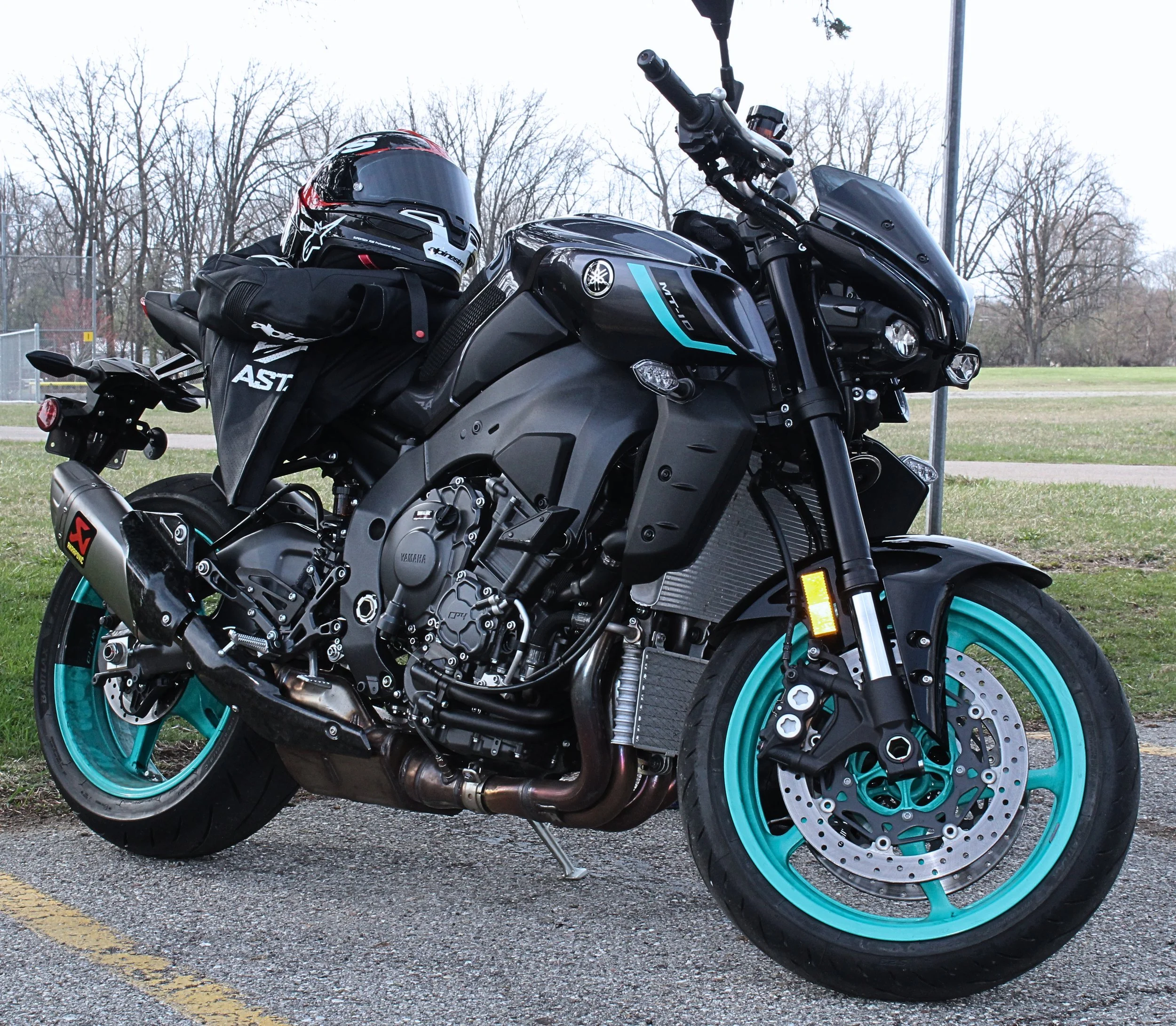 A black Yamaha motorcycle with teal accents parked outdoors on a gray asphalt surface, with a motorcycle helmet and riding gloves placed on the seat. The background shows leafless trees and a cloudy sky.