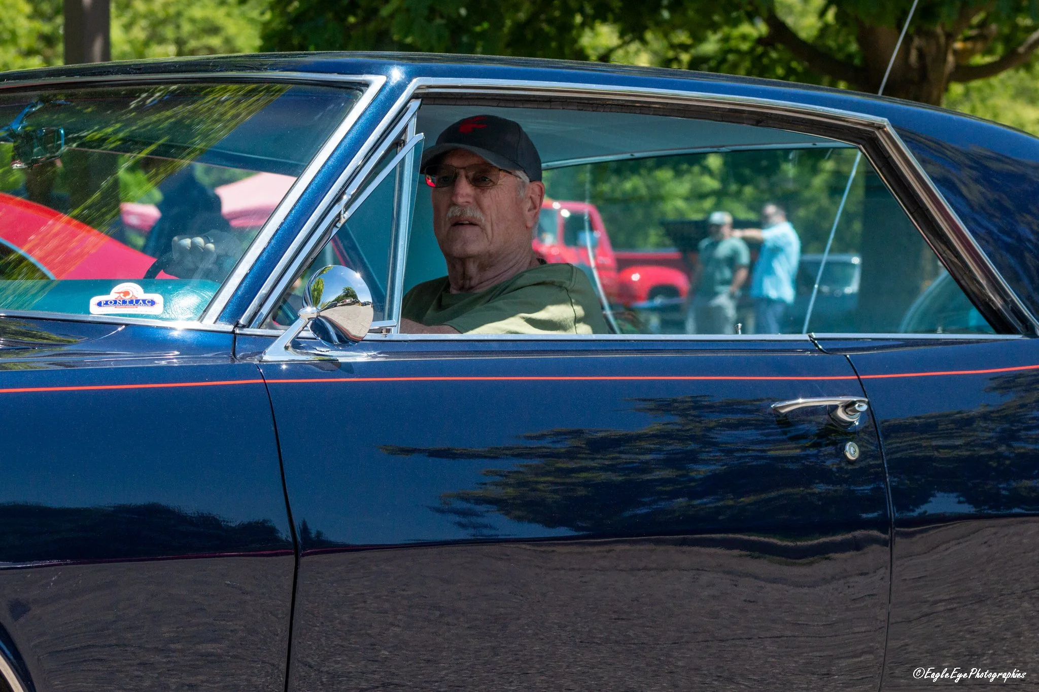 An older man wearing glasses and a baseball cap sitting inside a vintage navy blue car with a small sticker on the windshield that says 'Pontiac.' The car is outdoors with trees and people in the background.