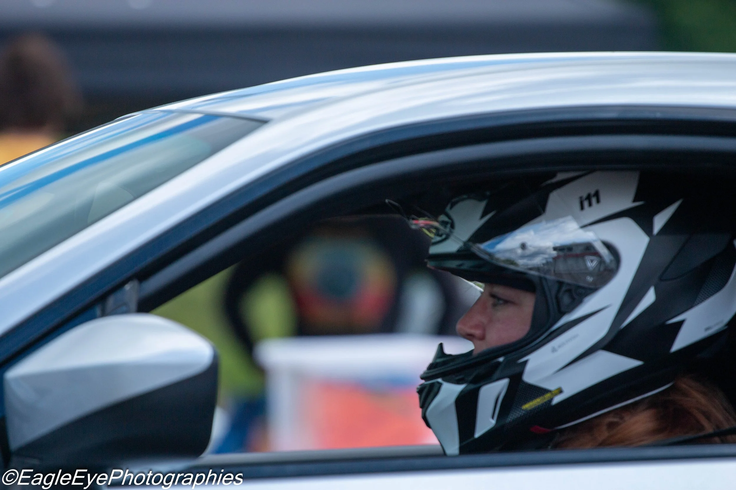 Close-up of a race car driver in a black and white helmet, sitting inside a race car with a white exterior. The driver appears focused, with a helmet strap fastened, and there are blurred background elements.