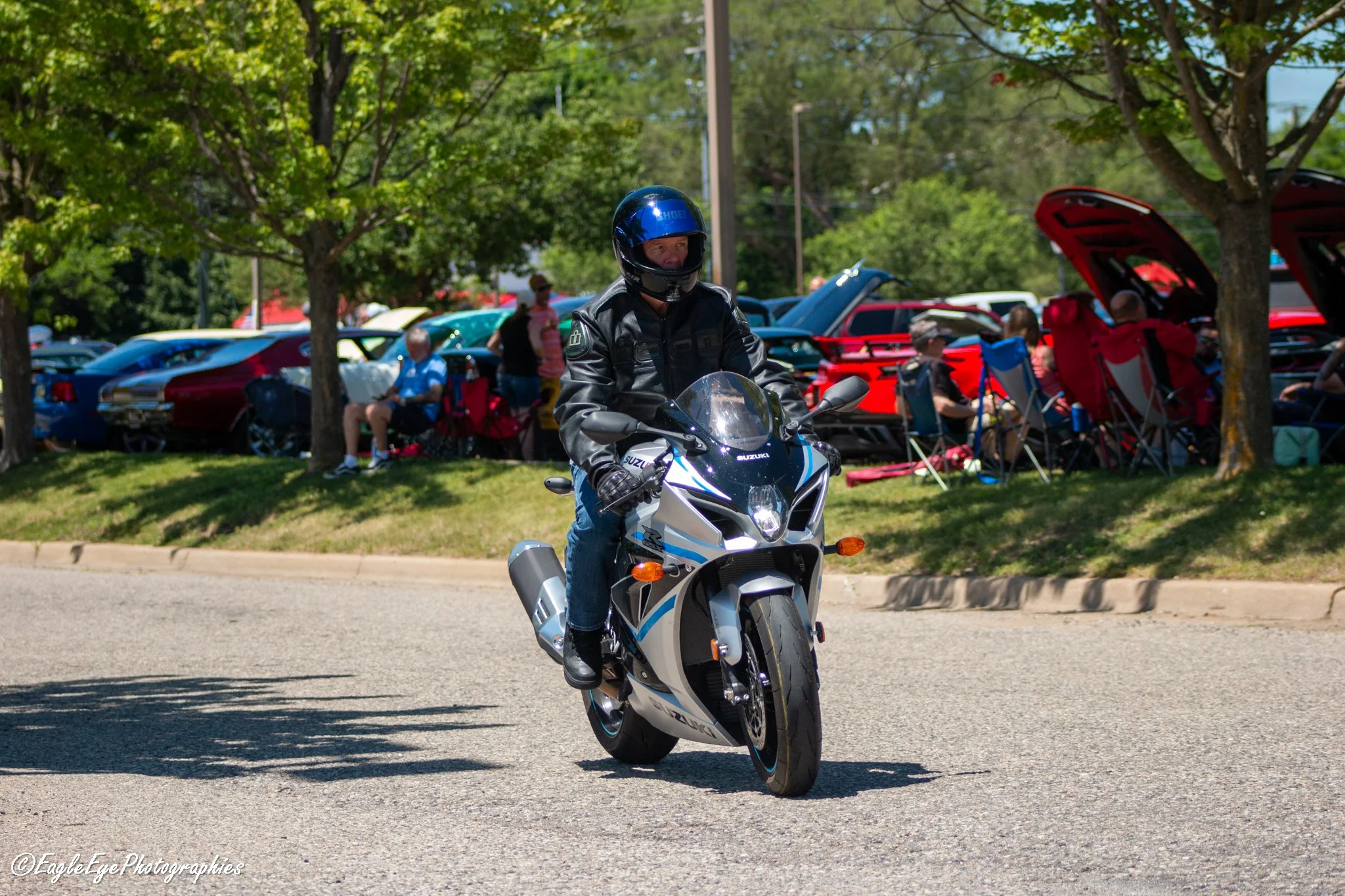 A person wearing a black helmet and leather jacket riding a Suzuki motorcycle on a street, with a backdrop of cars at a car show and people sitting in chairs under trees.