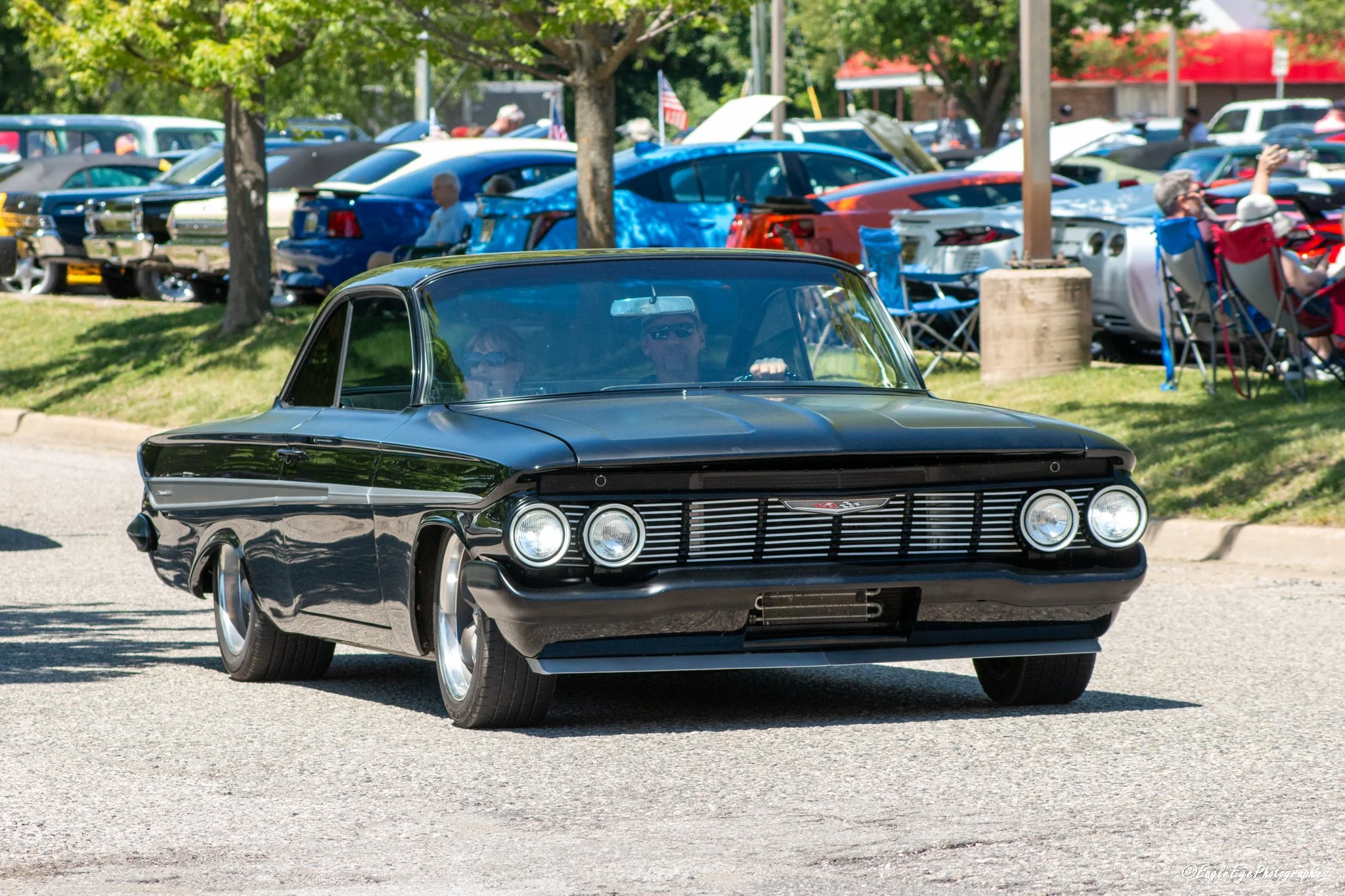 A black vintage car driving on a street with a car show in the background, featuring a white car with its hood open and tents with people in a park-like setting.