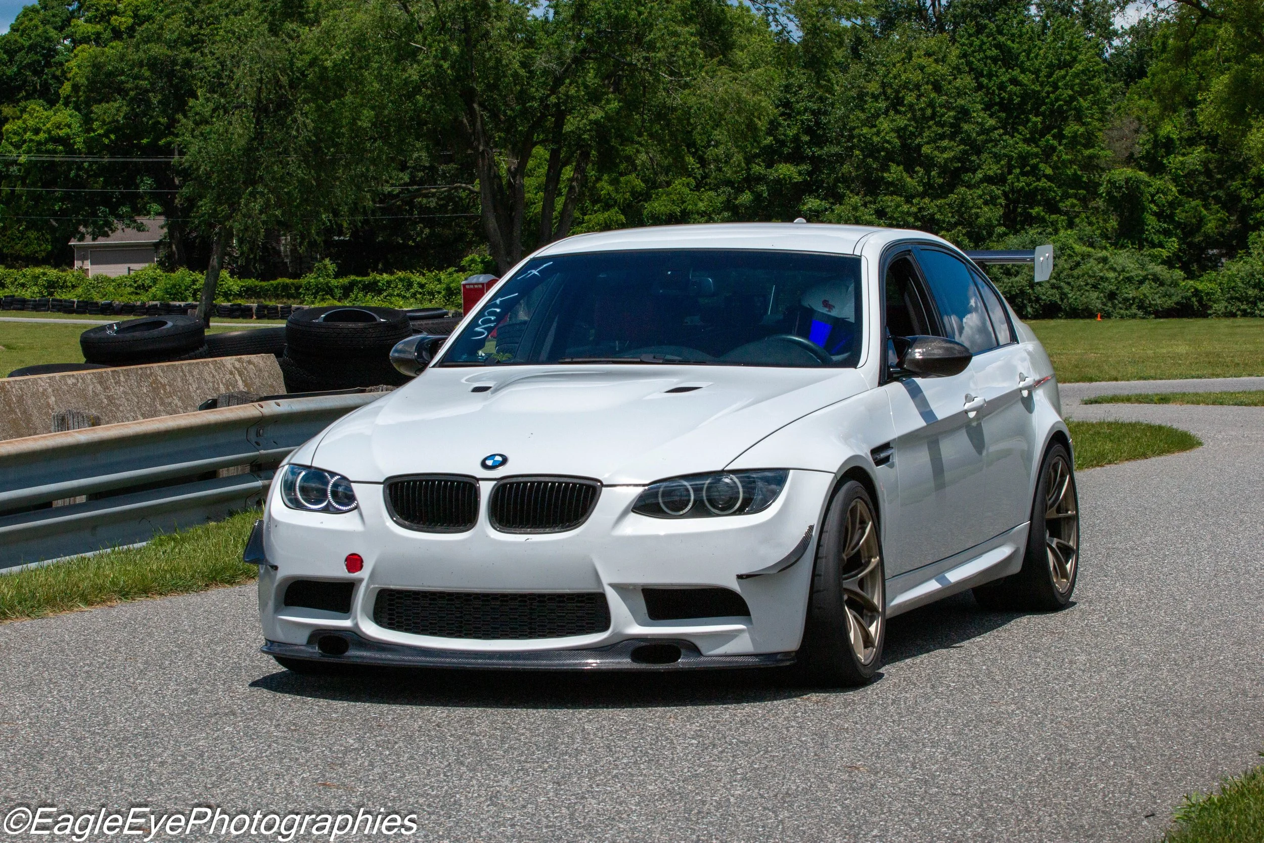 Close-up of a white BMW sports car parked on a paved road next to a guardrail, with trees and grassy area in the background, and tires stacked behind it.