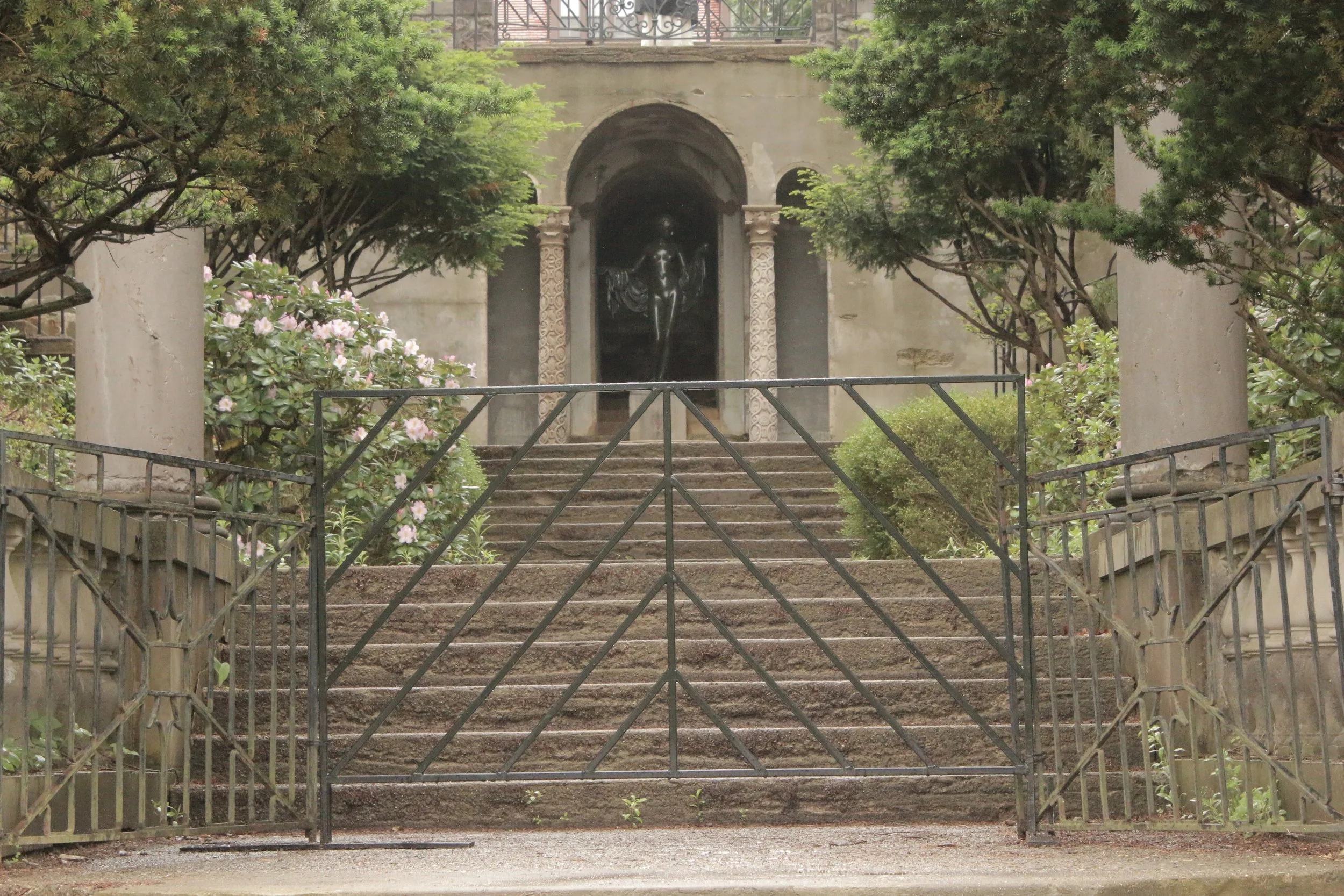 A set of stone stairs leading up to a gated entrance with decorative columns on either side. The stairs are framed by green bushes and trees, with a concrete wall and an archway with a statue inside at the top.