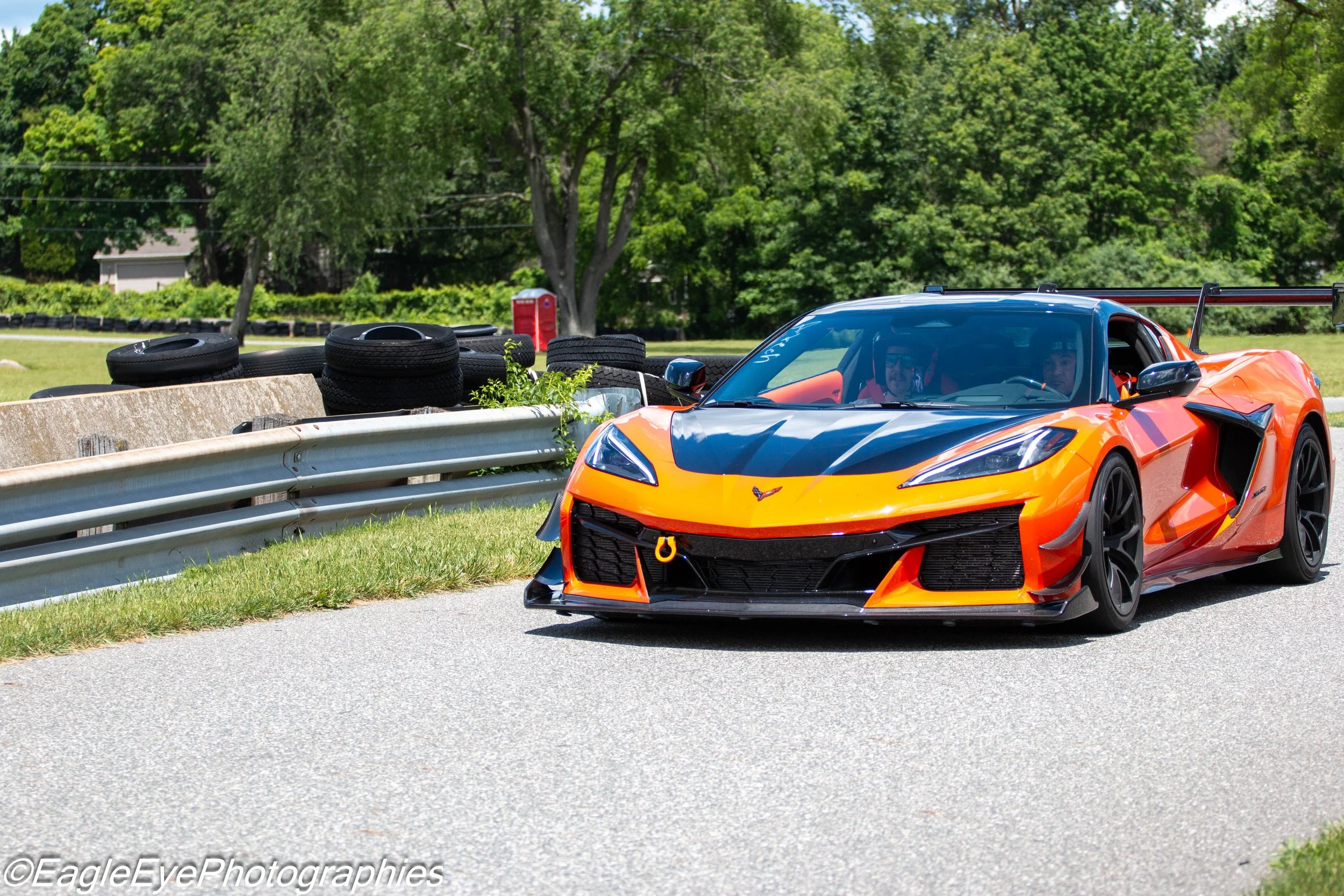 An orange and black sports car on a racetrack, with a driver and passenger inside, next to a barrier with tires and a red portable toilet in the background, surrounded by green trees.