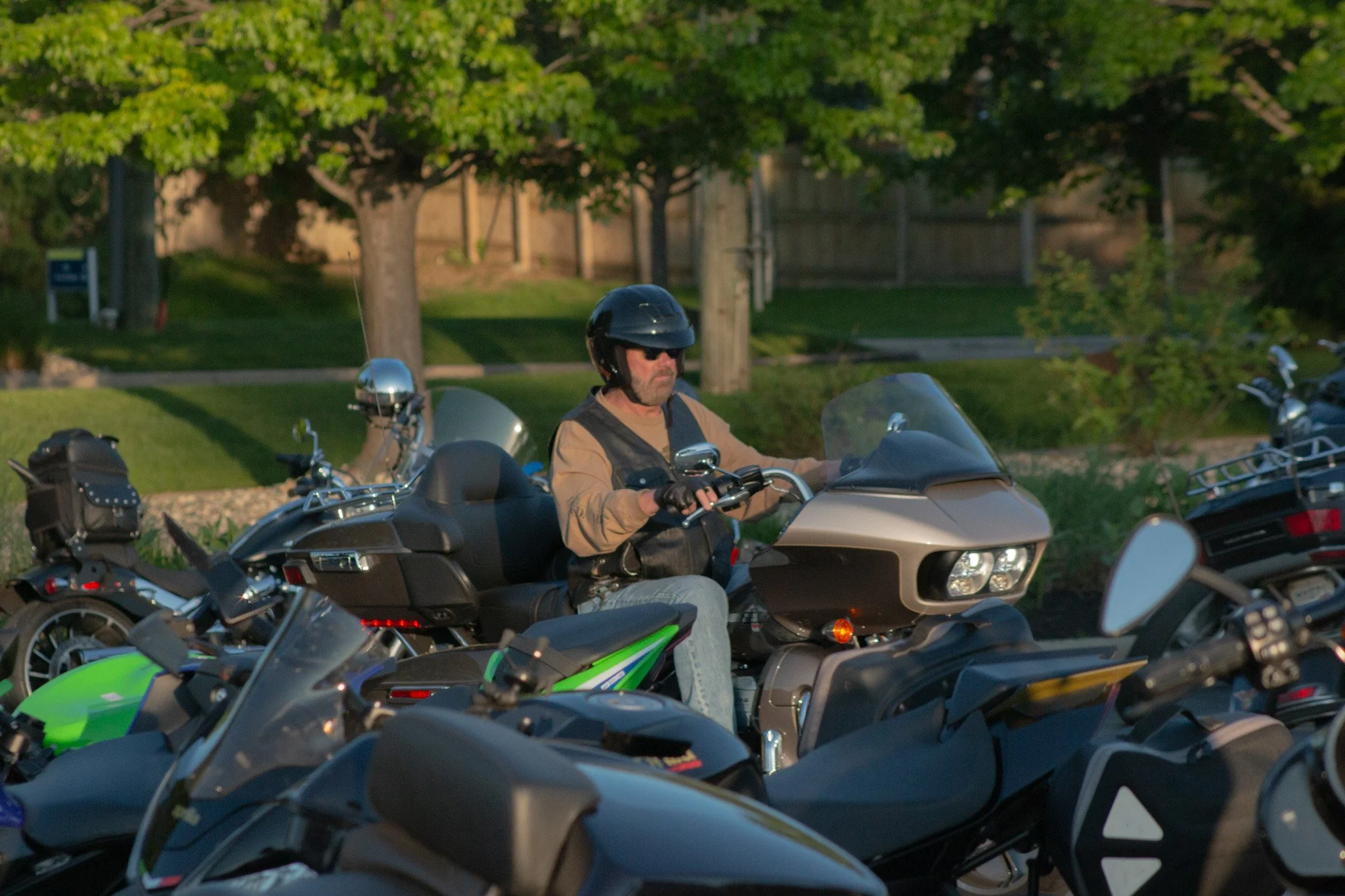 Man wearing a helmet and sunglasses riding a motorcycle among other motorcycles parked along a grassy area with trees and a fence in the background.