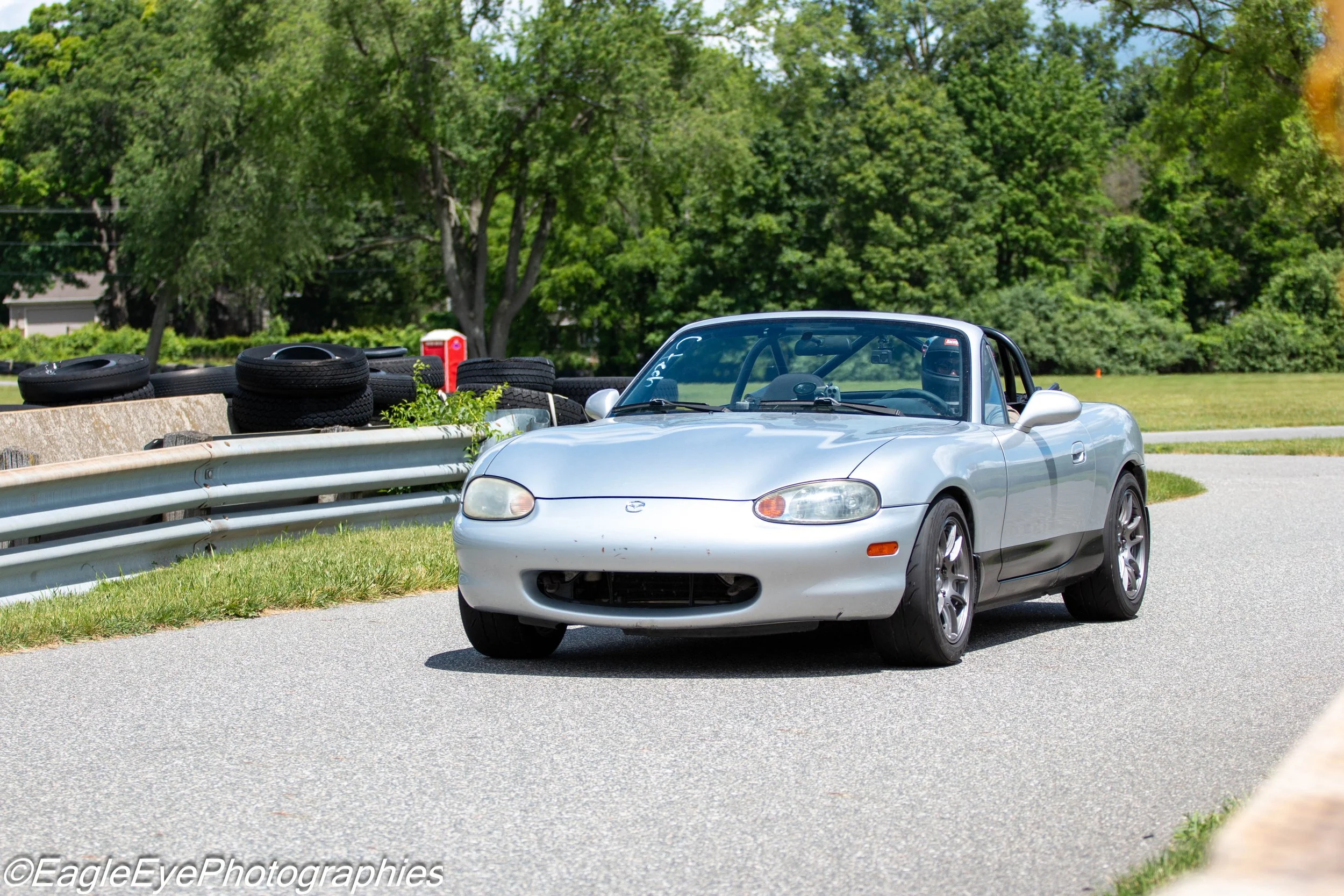 Silver convertible sports car parked on the side of a road with tires stacked on a barrier and green trees in the background.