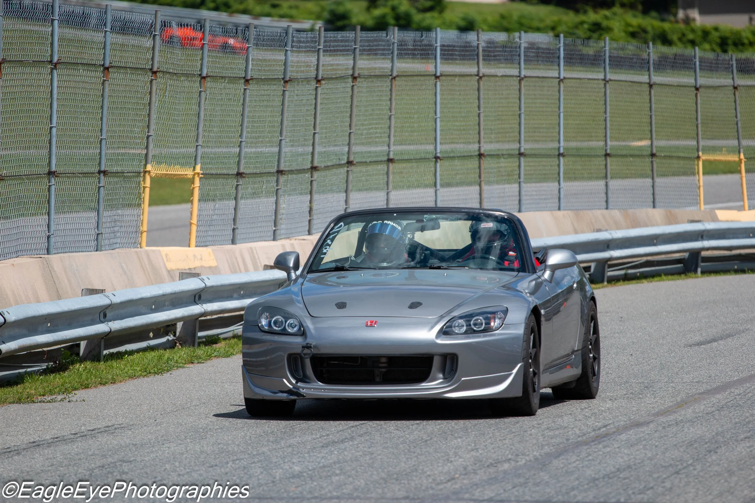 A silver sports car with a Honda emblem on the front, driven on a race track, with a fence and green trees in the background.