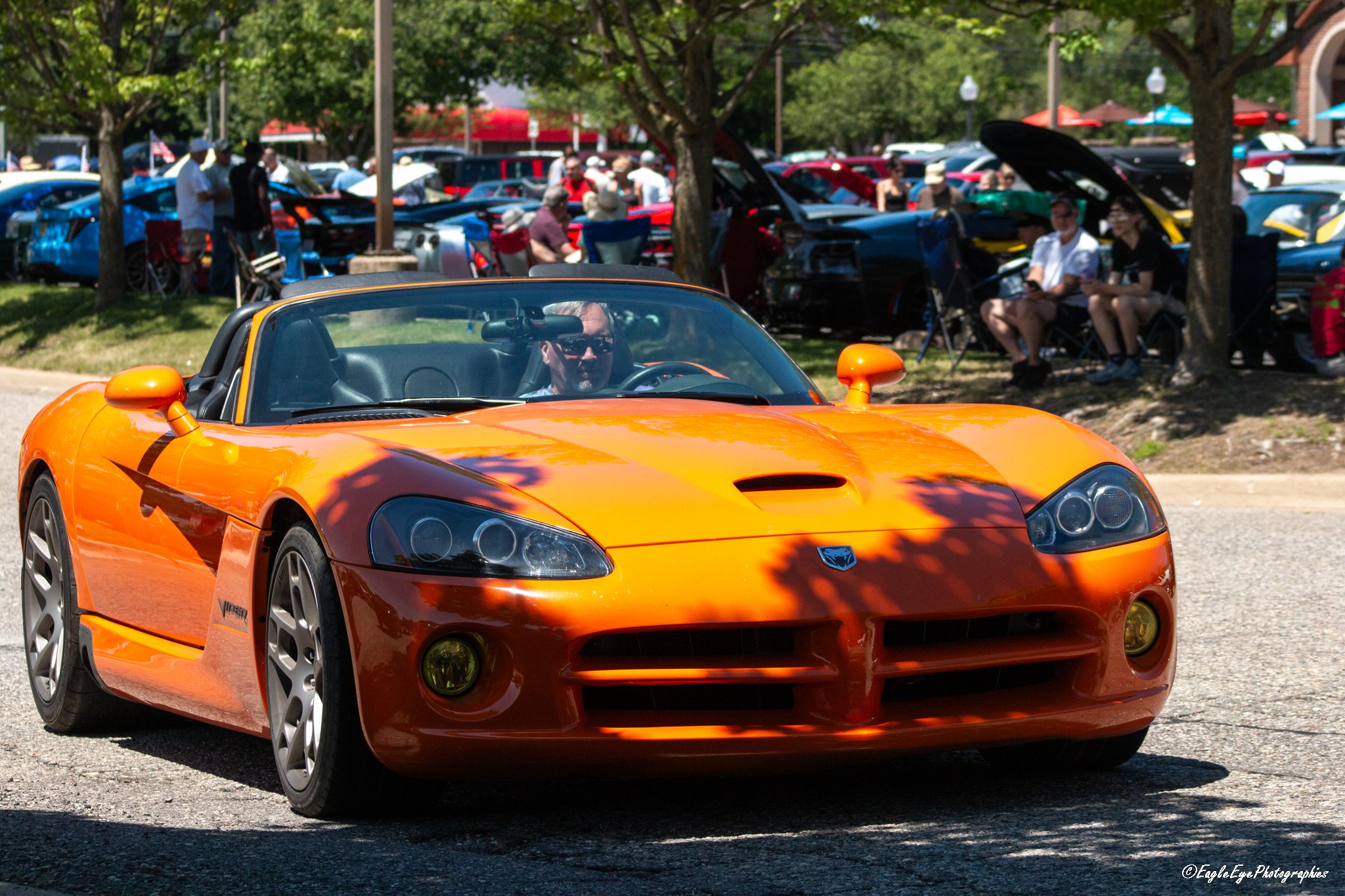 Orange sports car in the foreground with a man driving and multiple classic cars and people in the background at an outdoor car show.