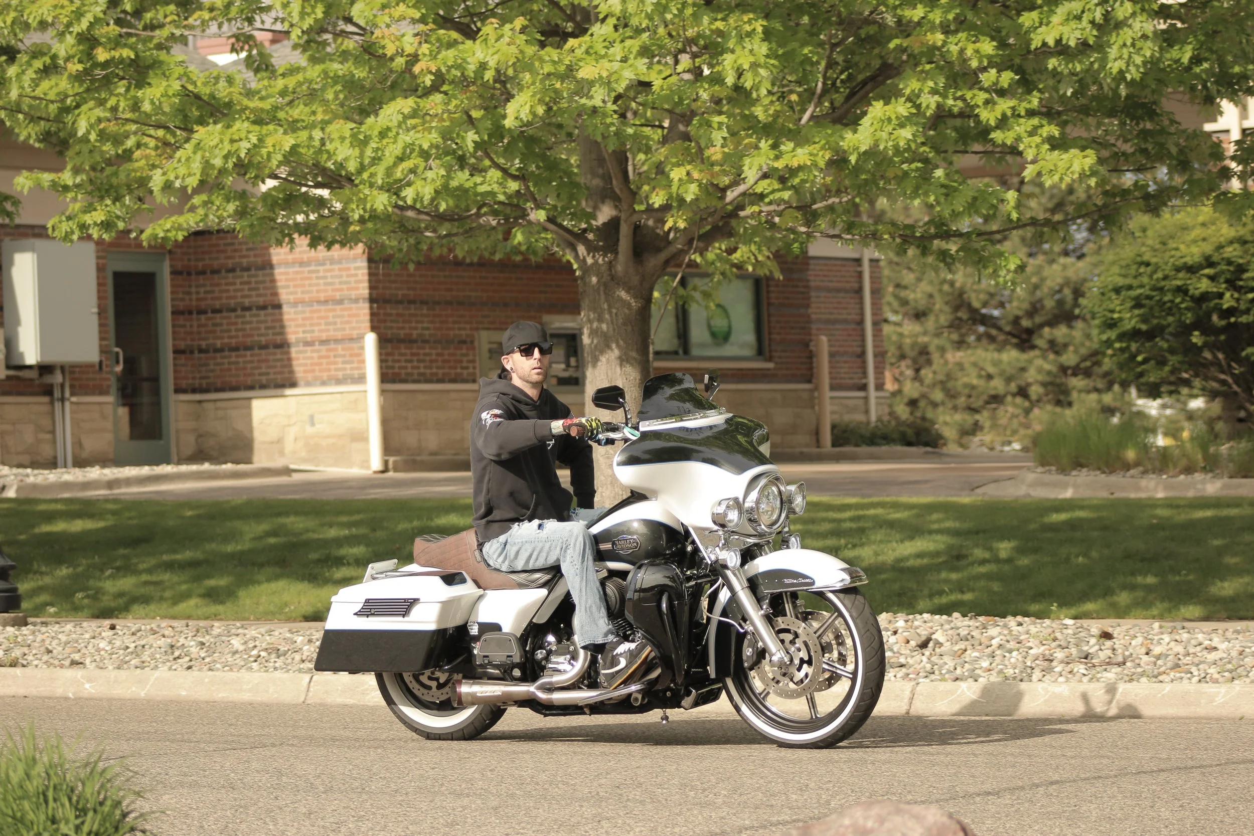 A man wearing sunglasses, a black hoodie, and jeans sitting on a white Harley Davidson motorcycle parked on the side of a street with a brick building and green trees in the background.