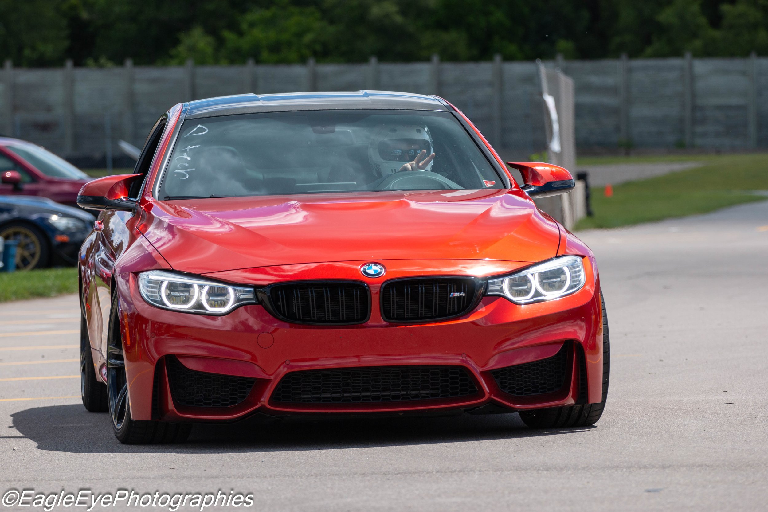Red BMW car parked on a lot with other vehicles, driver inside making a peace sign gesture, person wearing a helmet in the driver's seat.