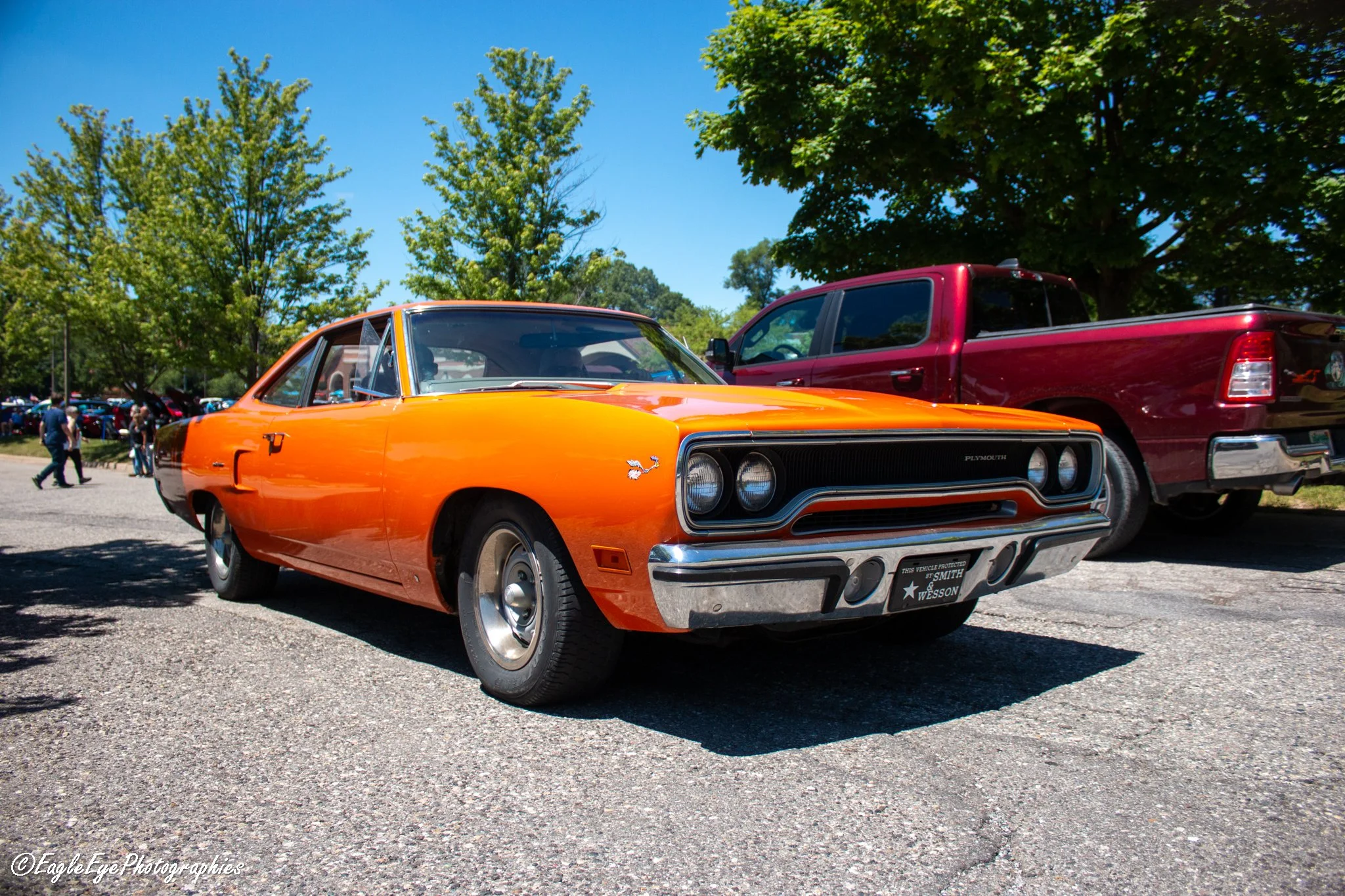 An orange vintage car, a Plymouth, parked outdoors on a gravel surface with a black front grill and chrome bumper, next to a red pickup truck, under trees with green leaves and a blue sky. People are walking in the background.