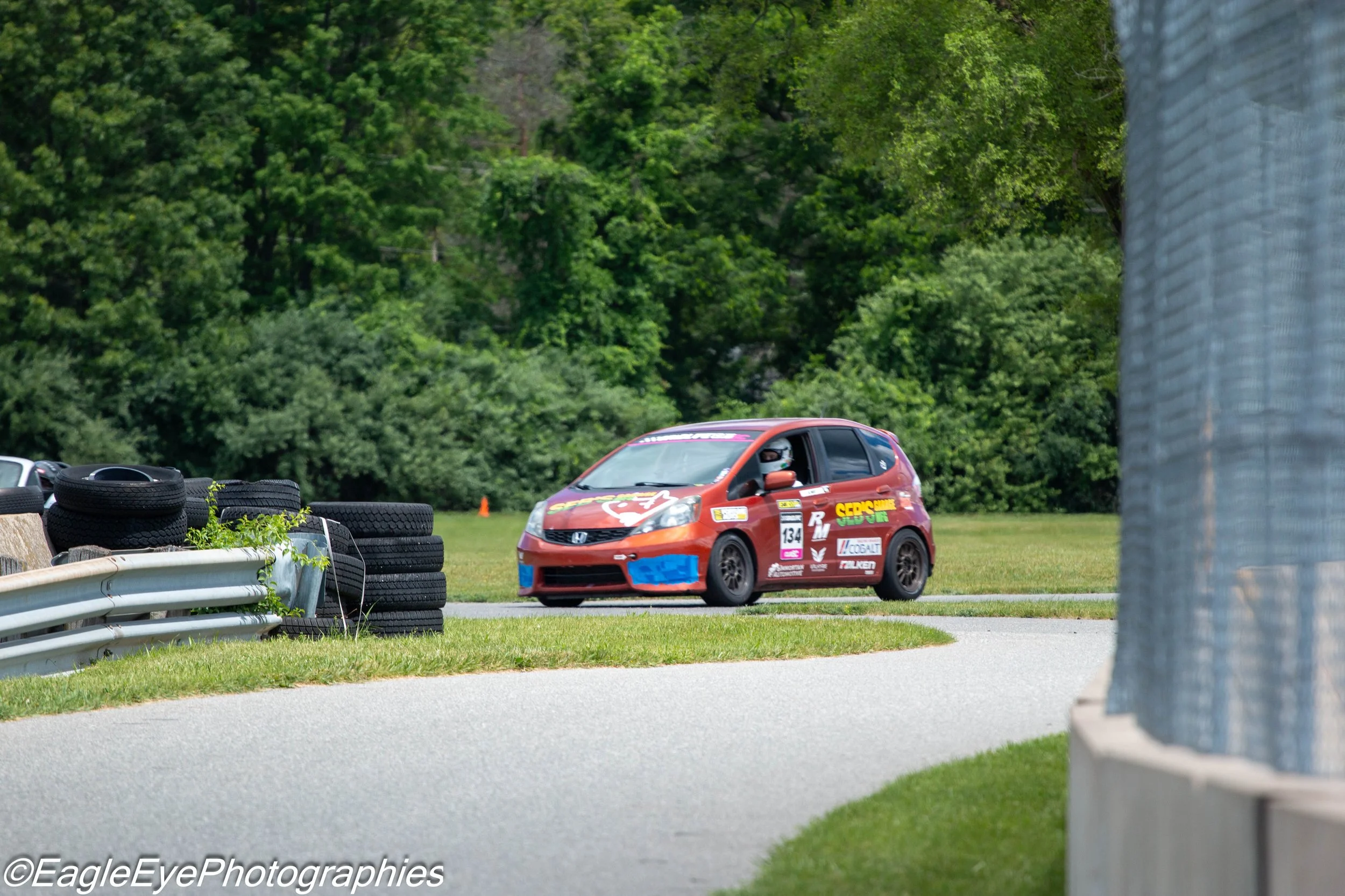 A small red race car with various sponsor logos, including 'SEB'S JUNK' and 'TALKEN,' is on a race track next to a stack of tires and a metal barrier. A driver wearing a helmet is visible in the car. The scene is surrounded by green trees and foliage