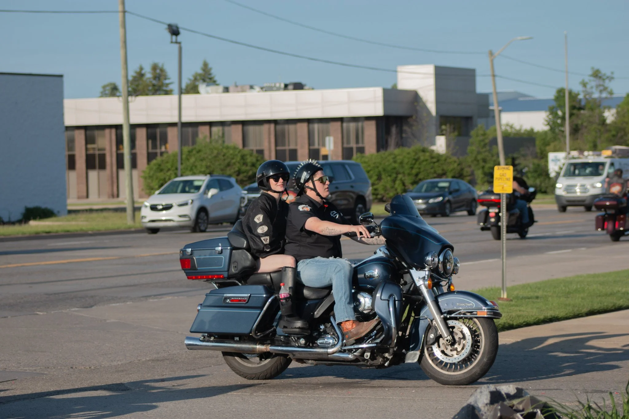 A man and woman riding a motorcycle on a city street, both wearing helmets and sunglasses. The man is driving, and the woman is sitting behind him.