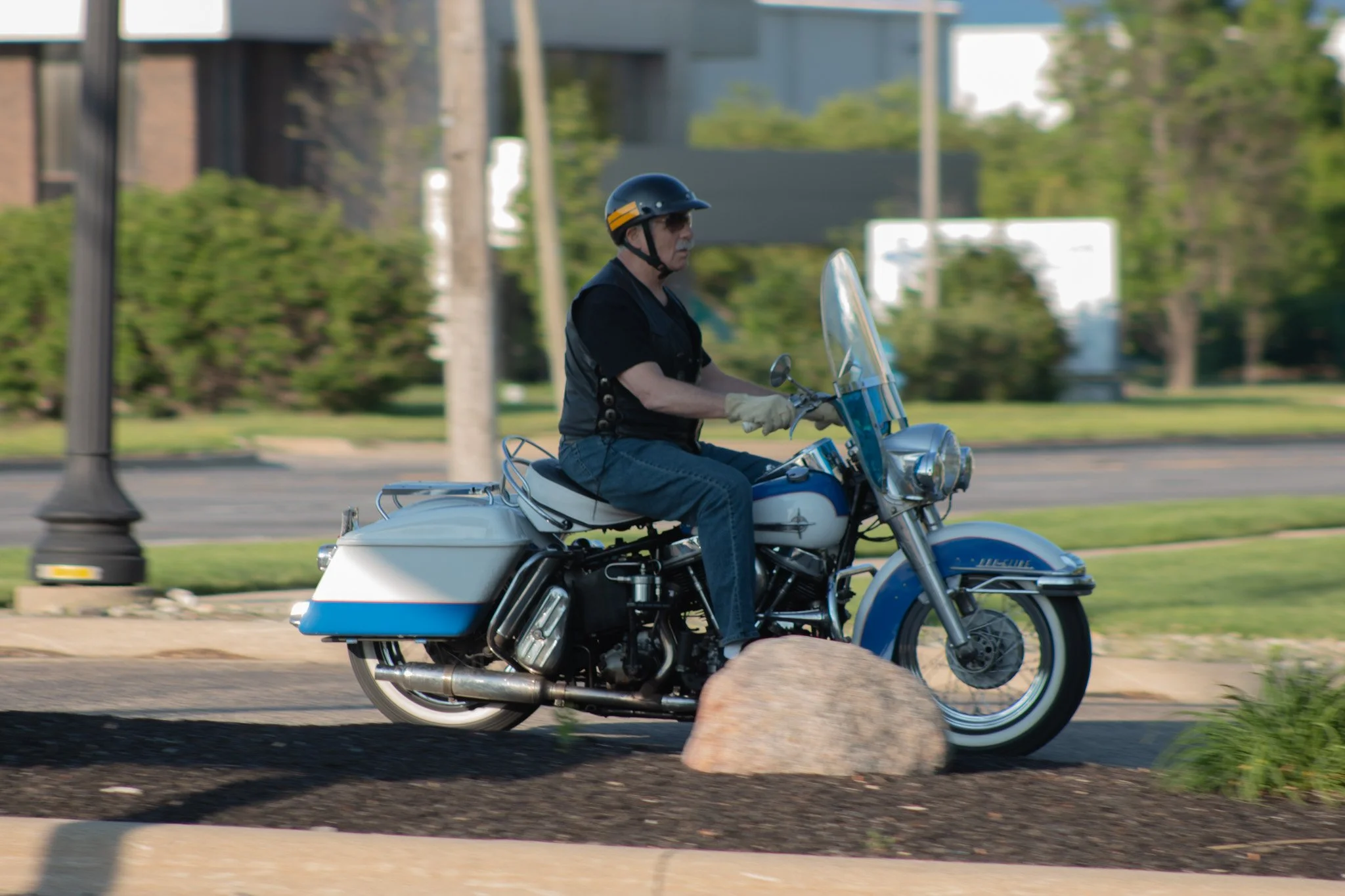 A man wearing a black helmet, sunglasses, gloves, and a black shirt riding a white motorcycle on a city street, with greenery and buildings in the background.