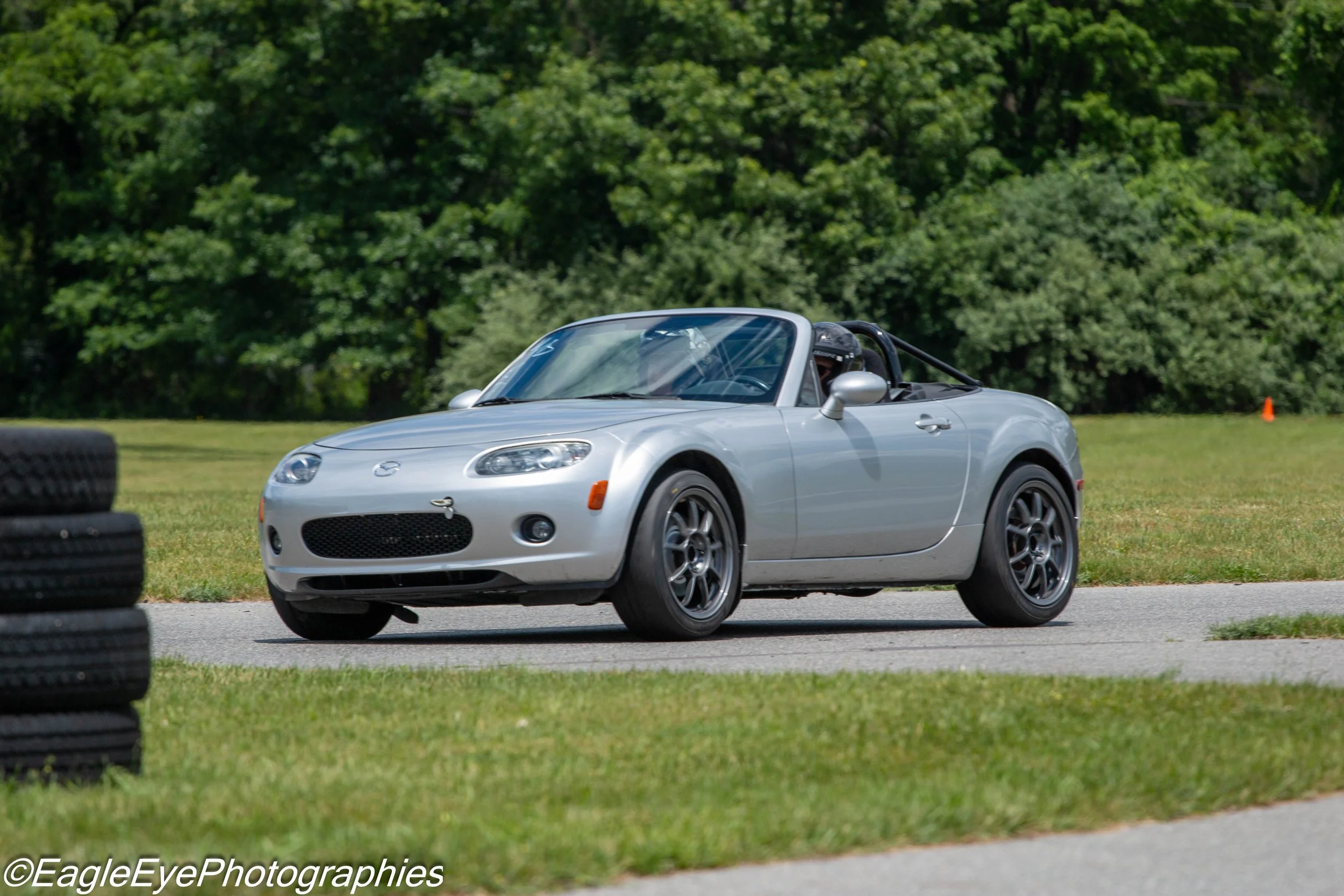 A silver Mazda MX-5 Miata convertible driving on a race track with green trees in the background.
