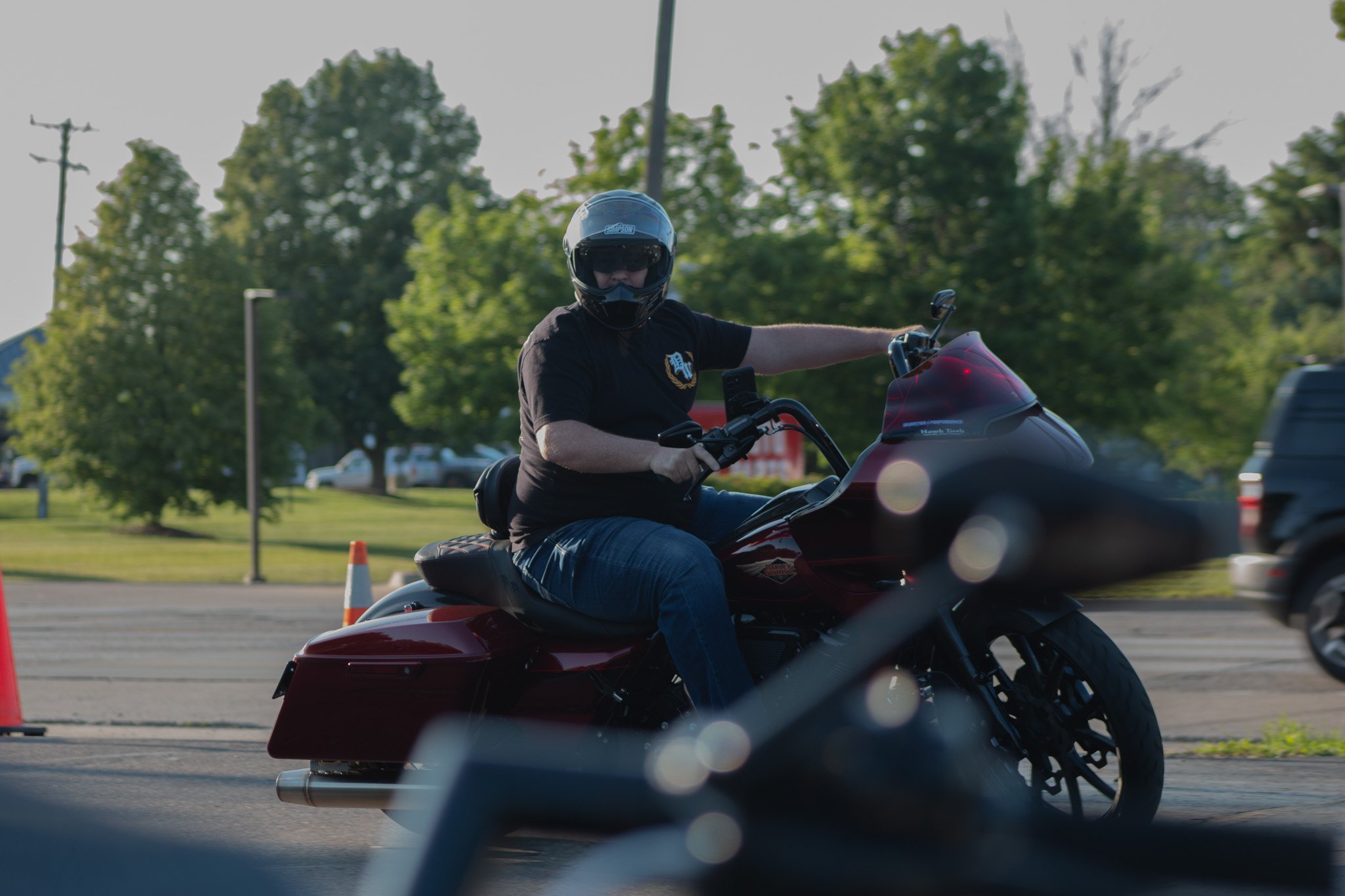 A person wearing a helmet riding a motorcycle at an outdoor event with trees and cars in the background.
