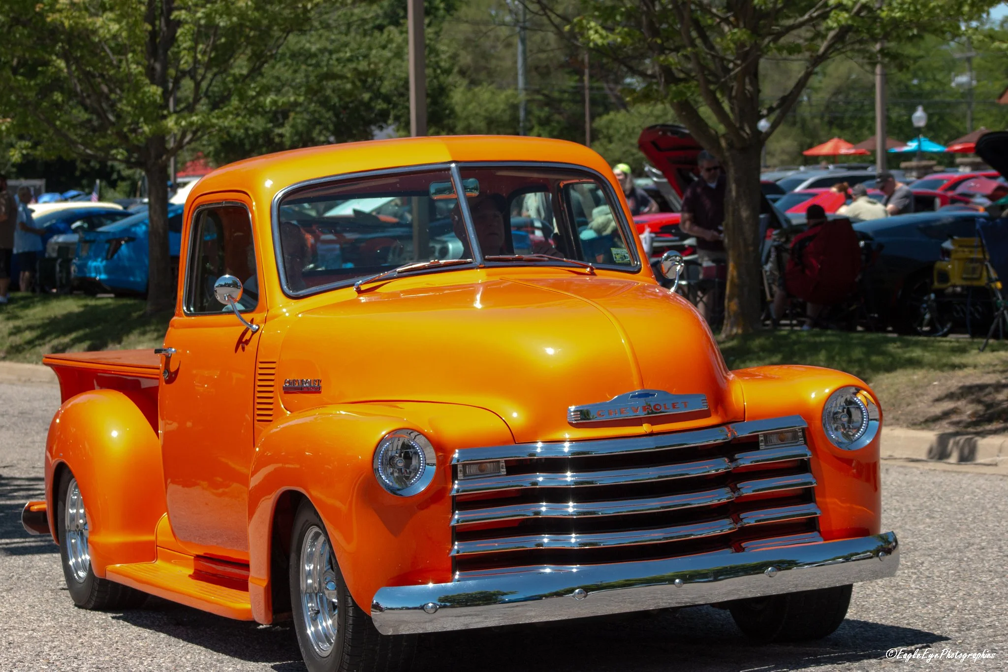 A shiny orange vintage Chevrolet pickup truck parked at an outdoor car show with other cars and trees in the background.