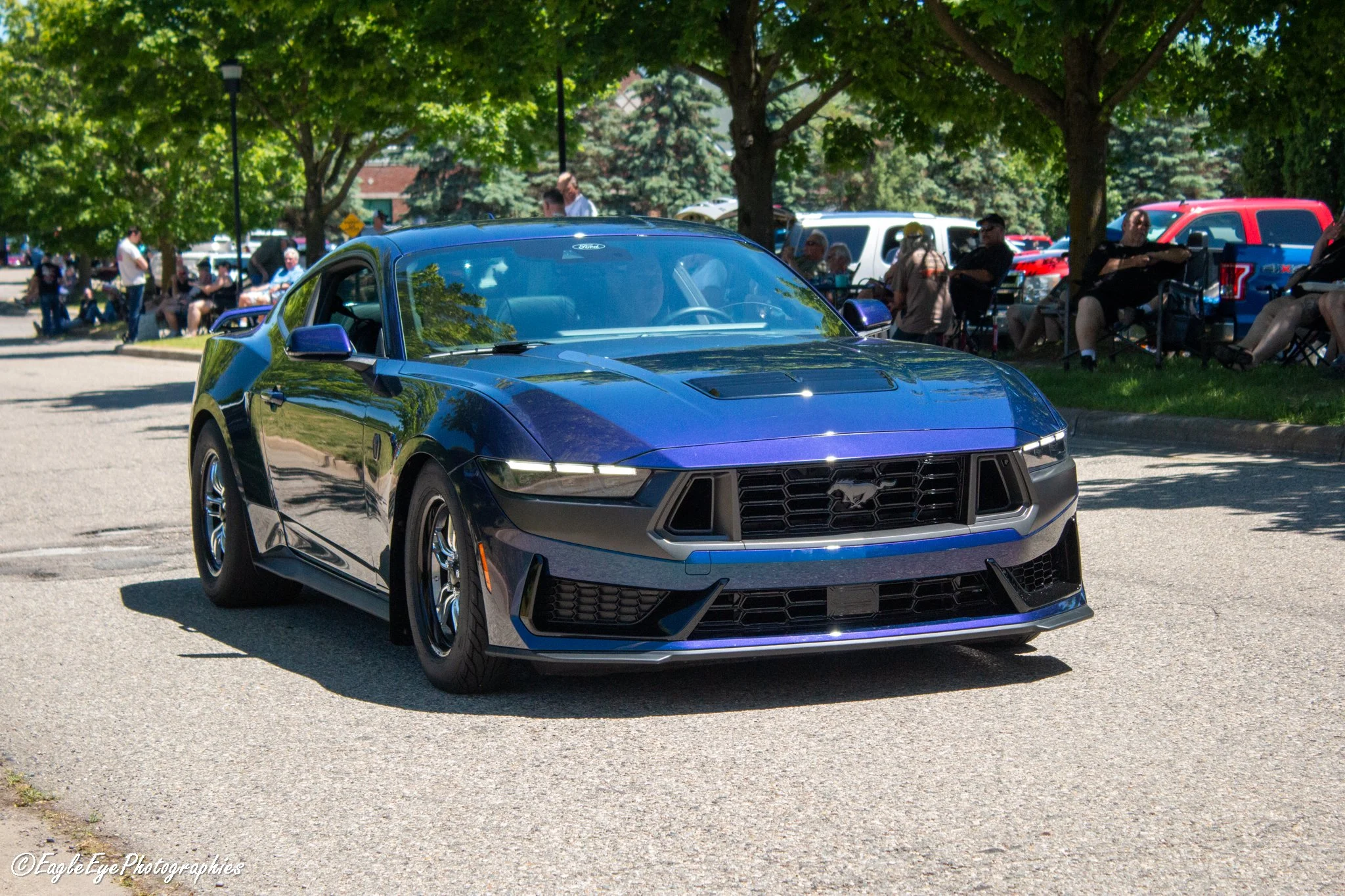 A blue Ford Mustang parked in a lot with several people sitting on benches and standing around under trees, some pickups and SUVs in the background, sunny day.