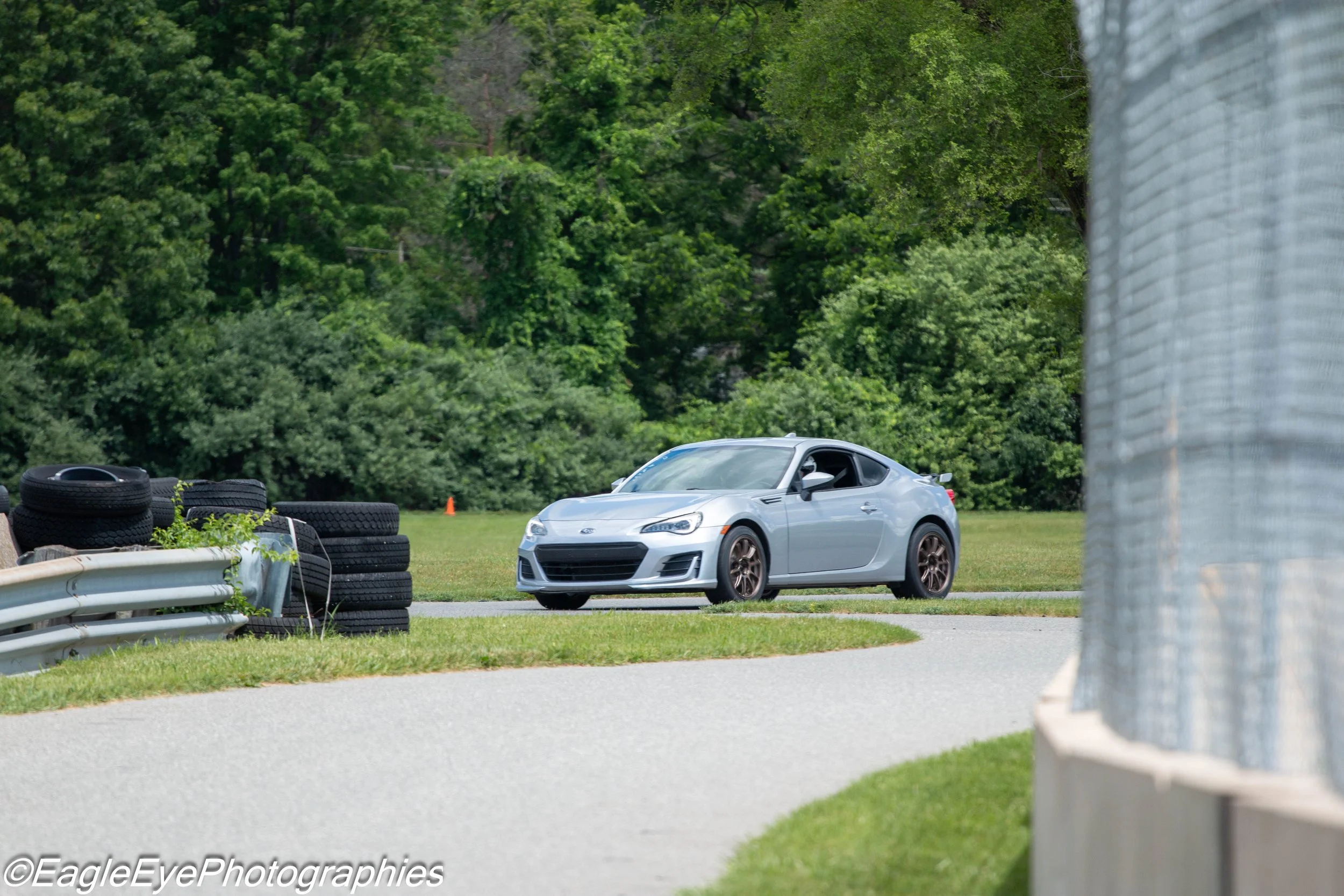 A silver sports car parked on a grassy area near a curved guardrail, with stacks of tires and a chain-link fence in the background, surrounded by lush green trees.
