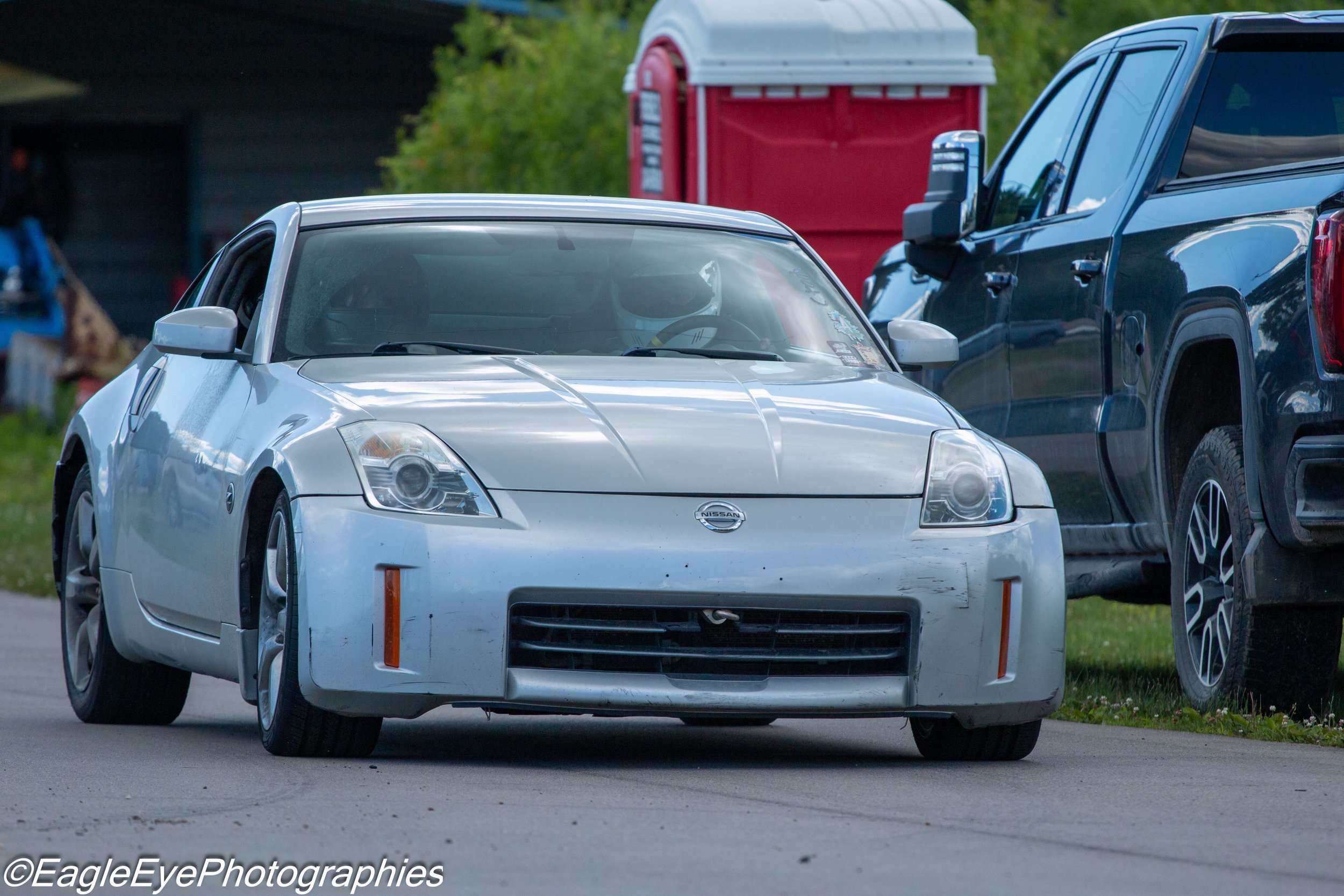 White Nissan sports car parked on a street, with a black pickup truck beside it and a red portable toilet in the background.