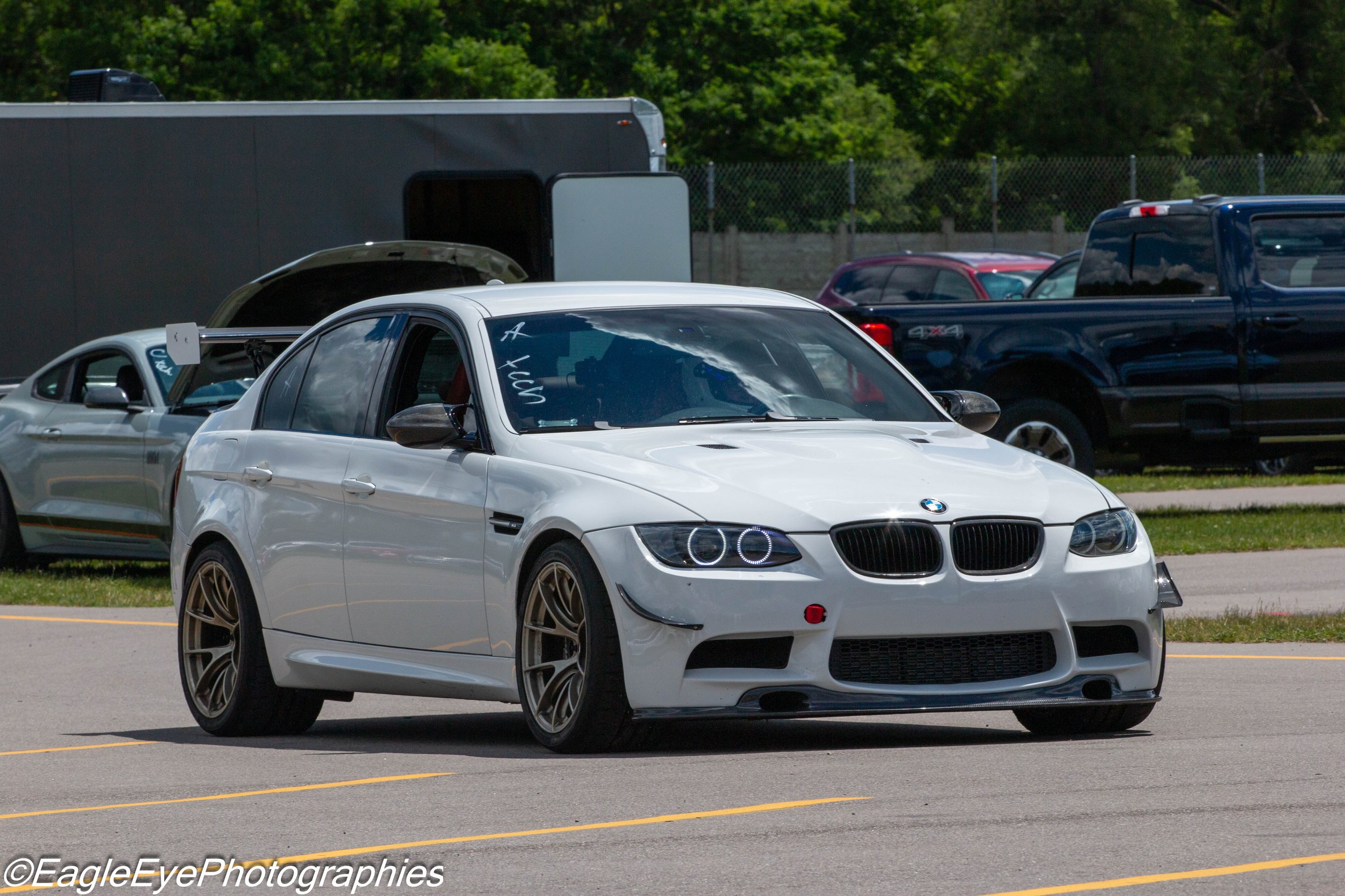 A white BMW sports car parked in a lot with other vehicles, including a gray trailer, a black truck, and a purple car, with a background of green trees and a chain-link fence.