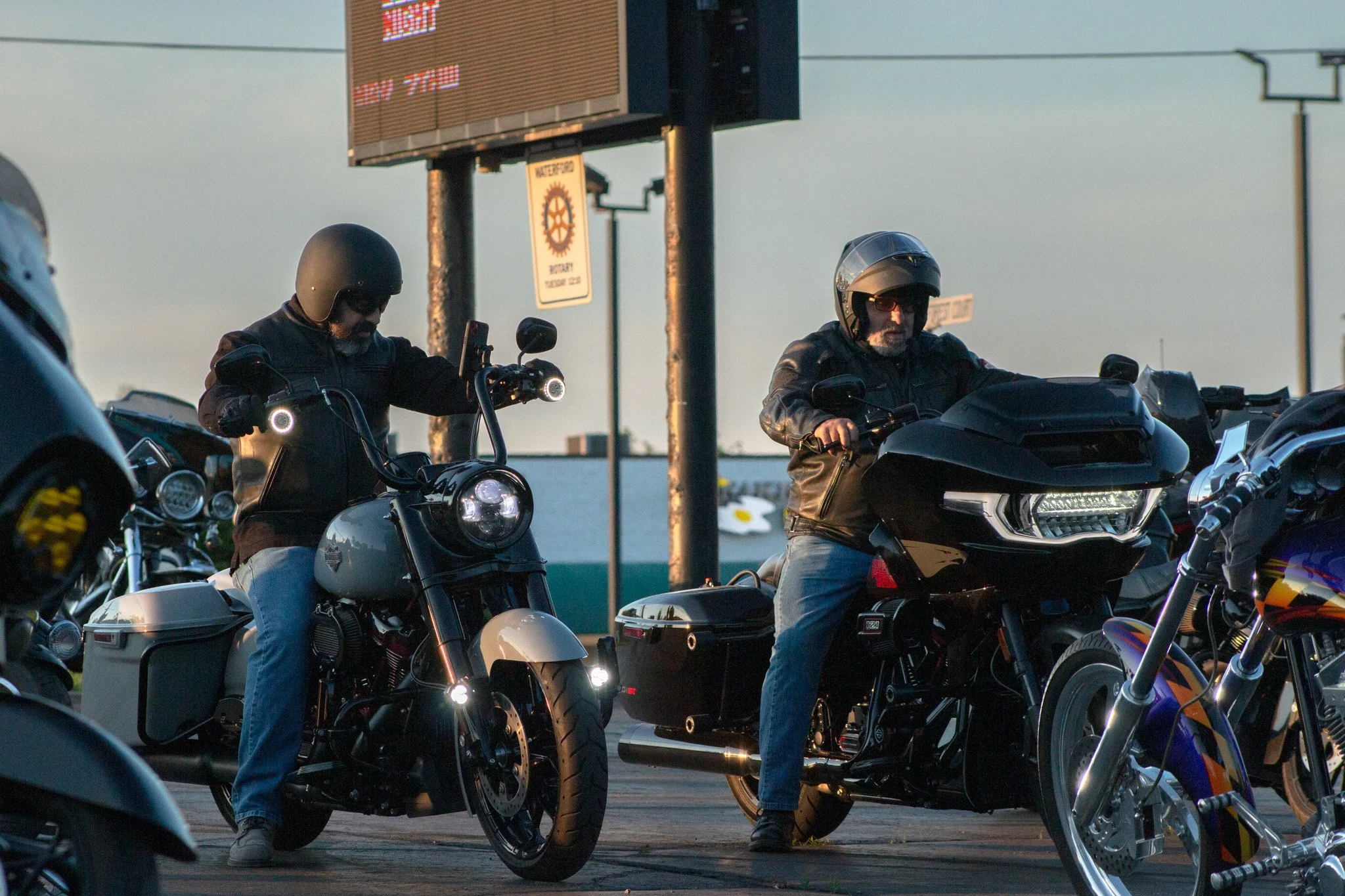 Motorcycle riders wearing helmets sitting on their bikes in a parking lot at sunset.