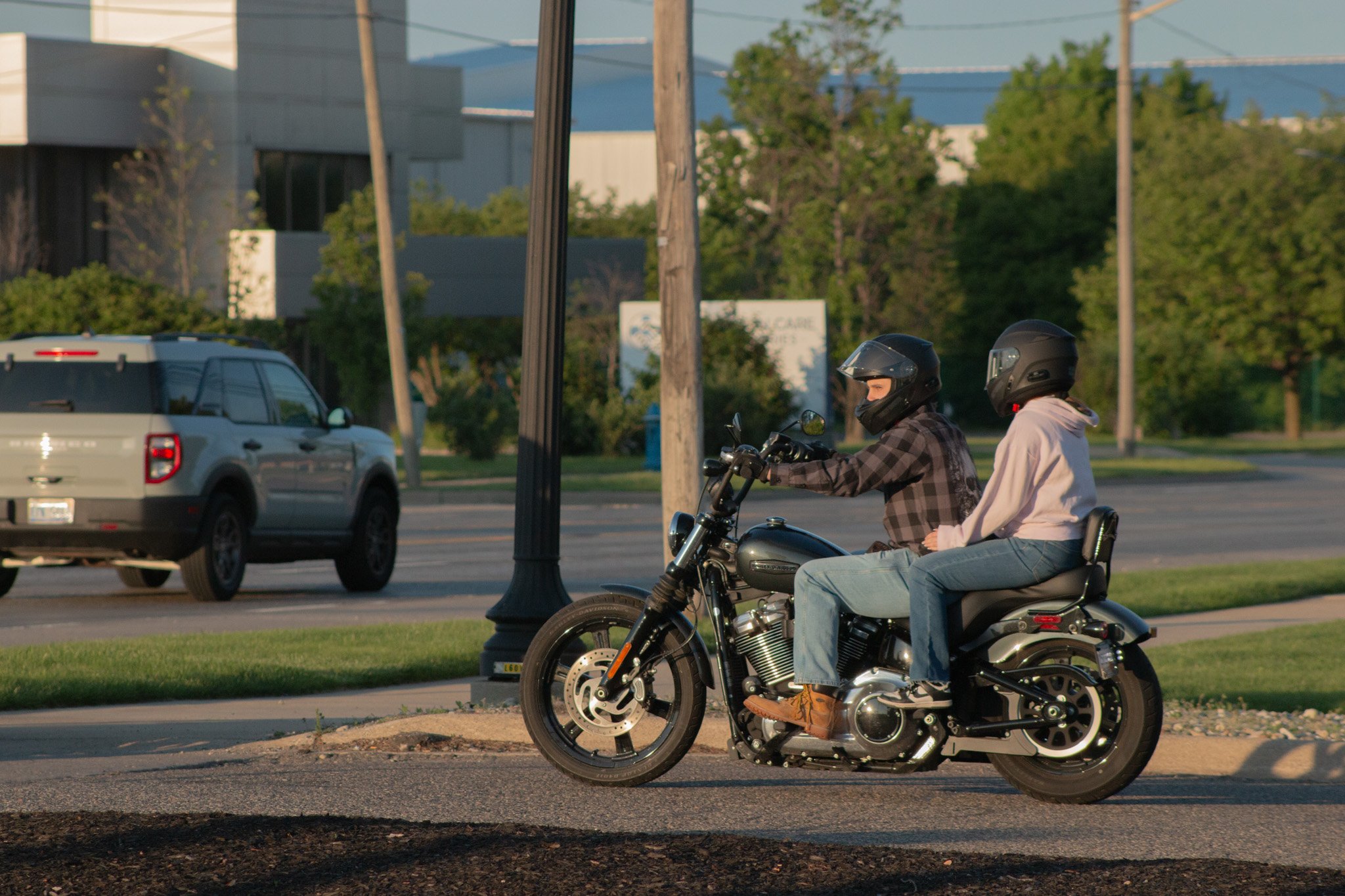 Two people wearing motorcycle helmets sitting on a motorcycle parked on the side of the road, with cars and trees in the background during daytime.