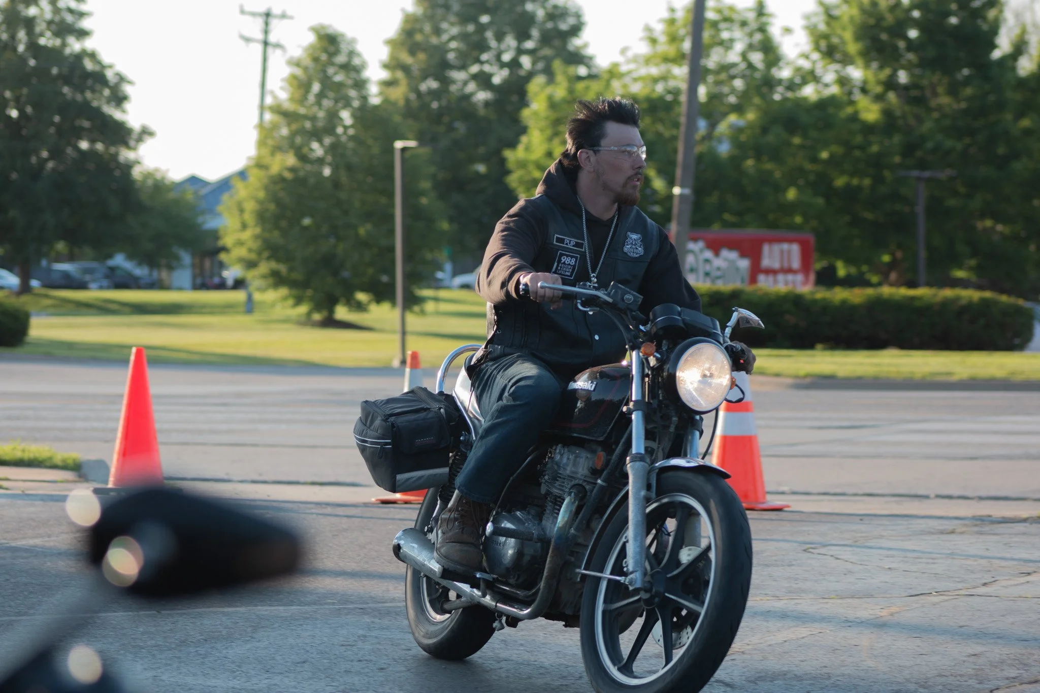 A man riding a motorcycle through an obstacle course marked by orange traffic cones outdoors. He is wearing a black jacket, glasses, and has a short beard and styled black hair. The background includes green trees and a sign that reads "AUTO".