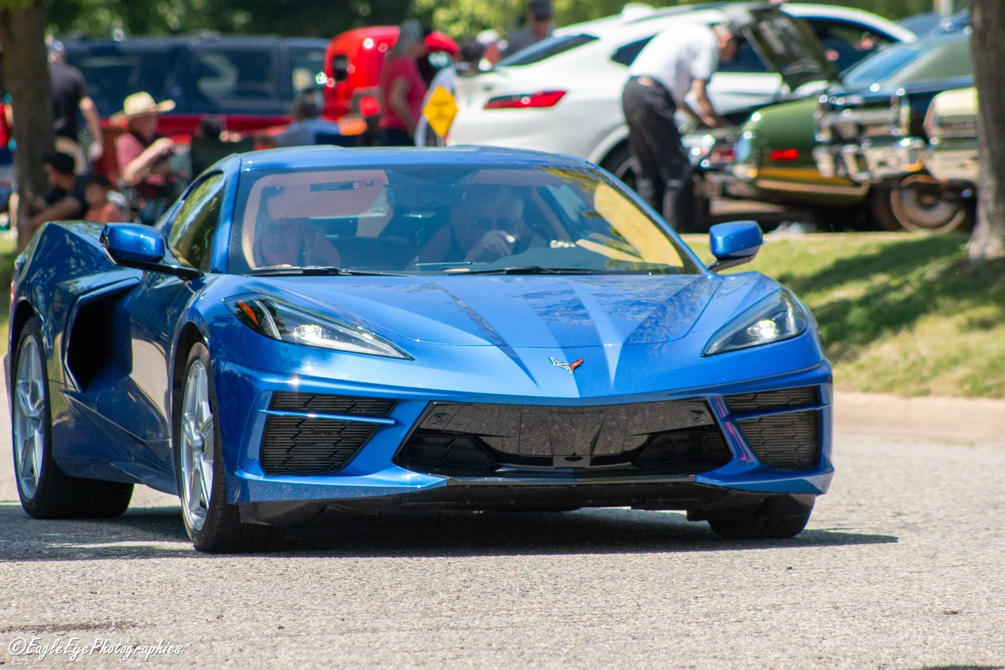Blue sports car in the foreground with people and other cars in the background at an outdoor event.