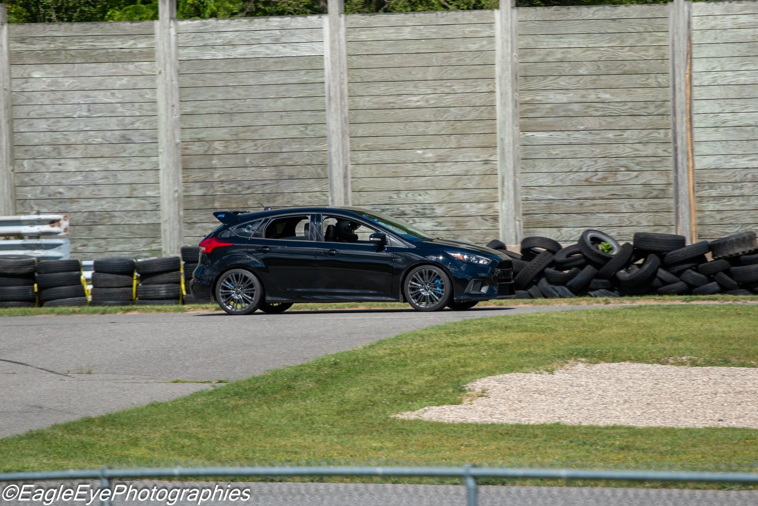 A black sporty hatchback car on a racetrack with a wooden barrier and tire barriers in the background.