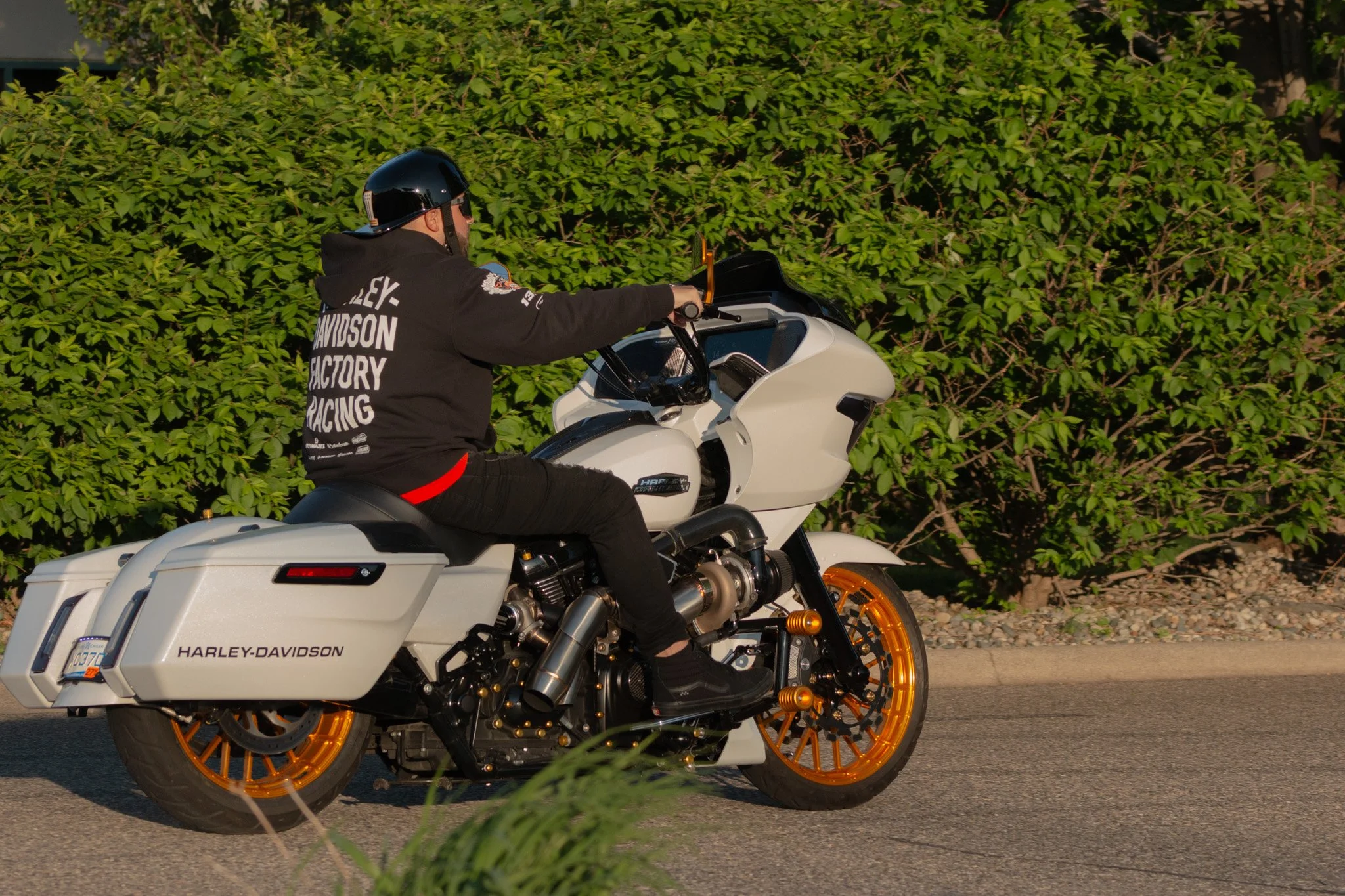 A person wearing a black Harley-Davidson hoodie and a black helmet riding a white motorcycle with orange wheels on a street with green bushes in the background.
