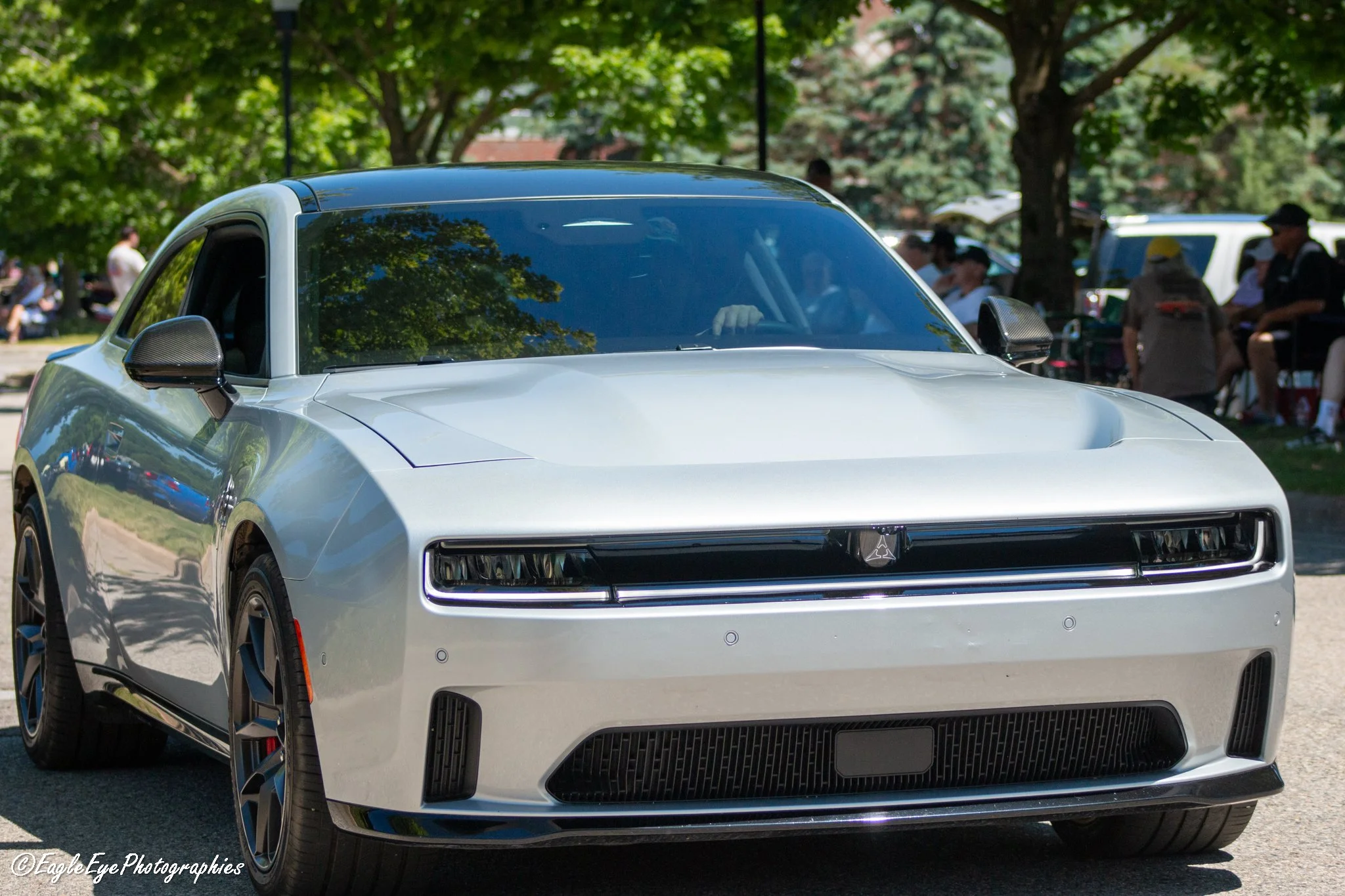 Front view of a silver electric car with a sleek design, black grille, and partially visible headlights, parked outdoors on a sunny day with trees and people in the background.
