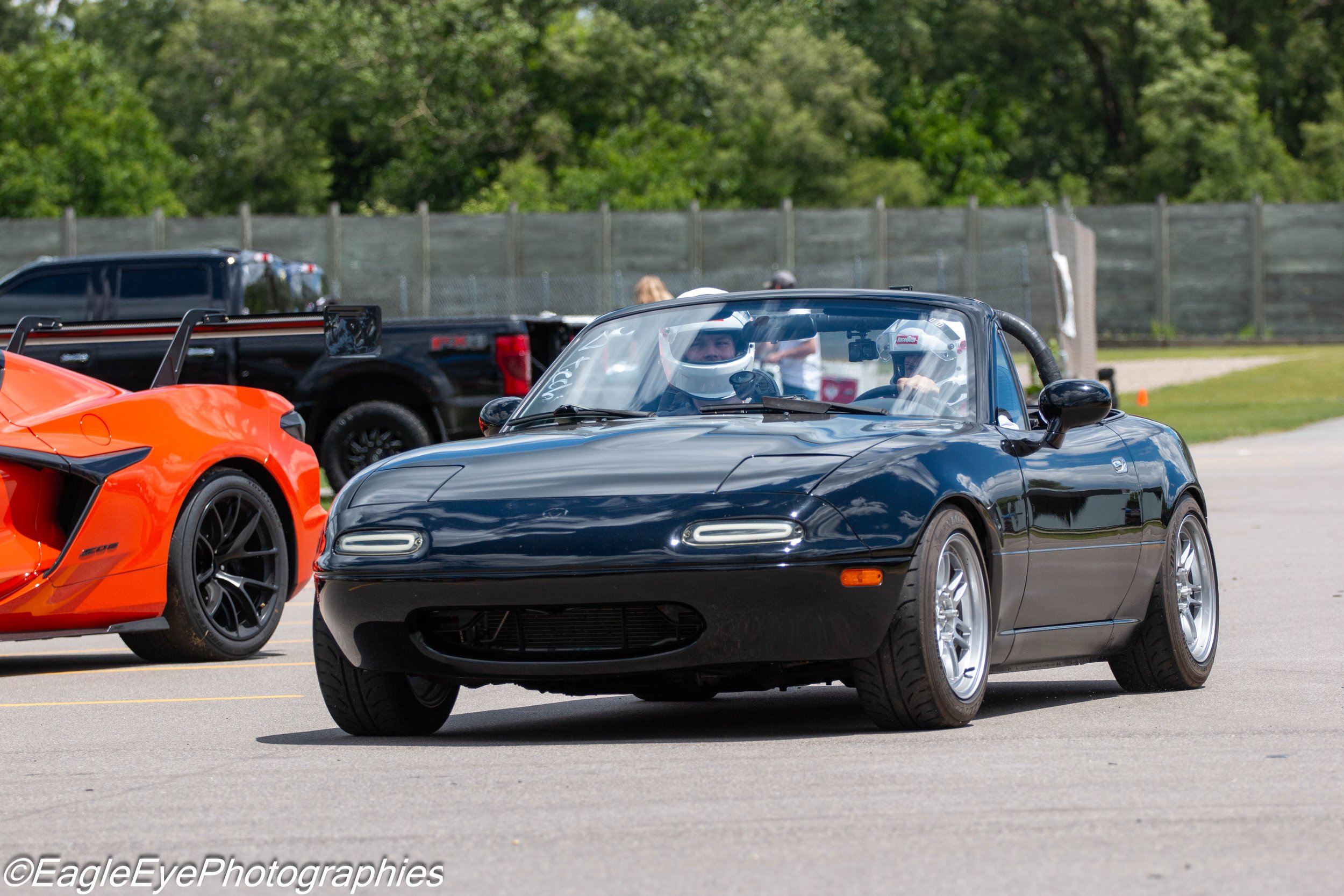 A black Mazda MX-5 Miata sports car with a woman driver wearing a helmet, parked on a paved lot during a car event, with a red sports car to the left and a black pickup truck in the background.