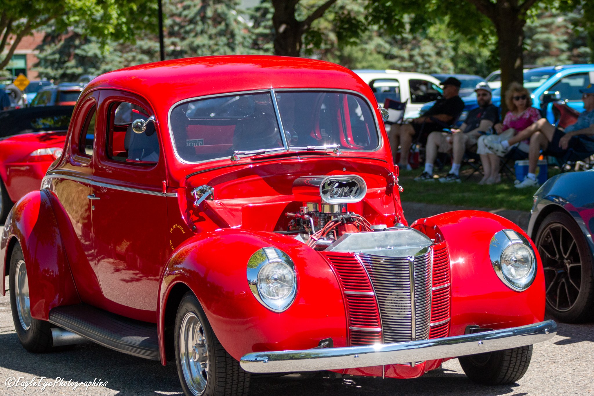A vintage red car on display at an outdoor car show with trees and people sitting in the background.