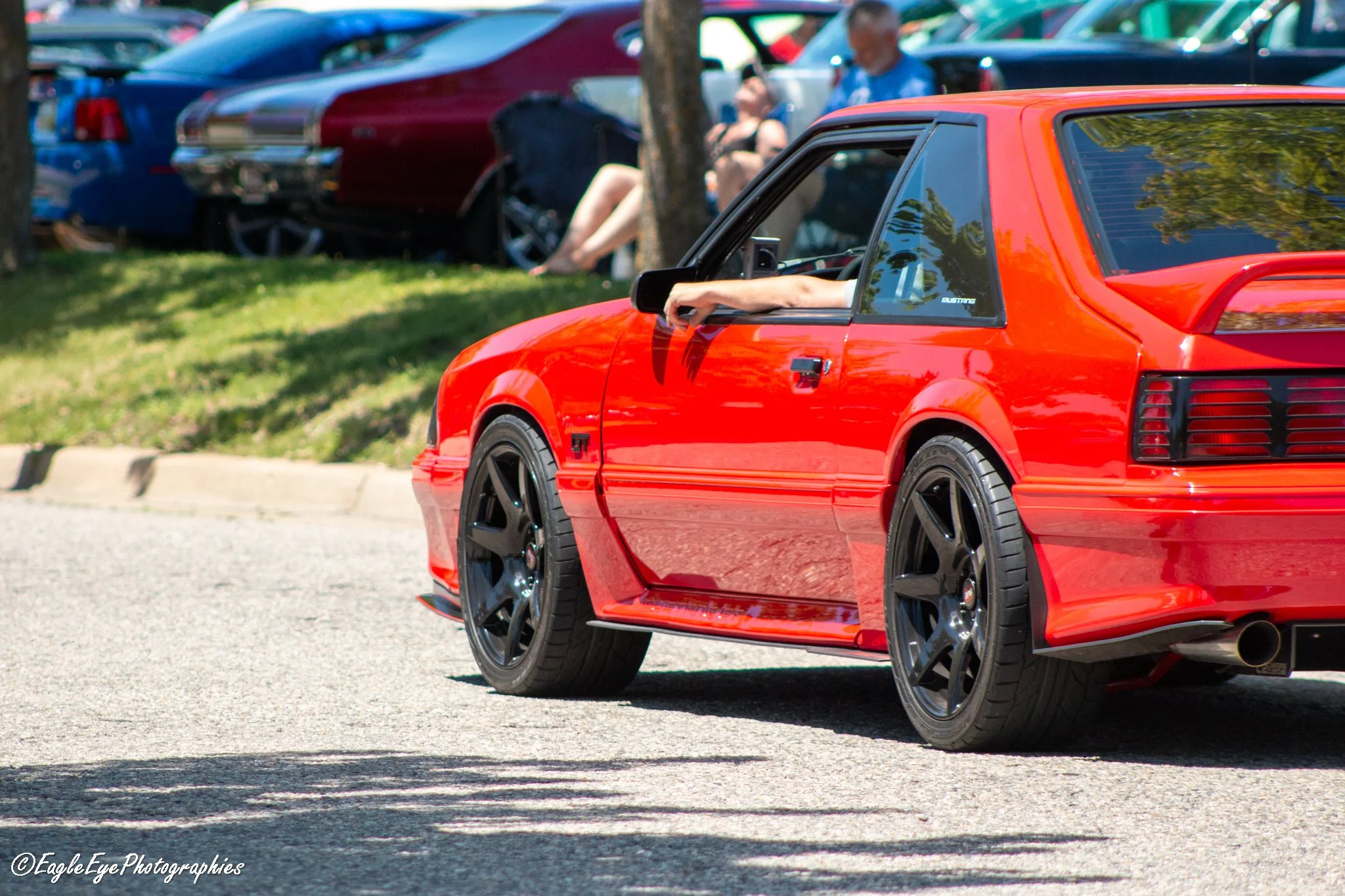 A red sports car parked on a street in front of a grassy area with trees, people sitting and standing, and other vehicles parked nearby.